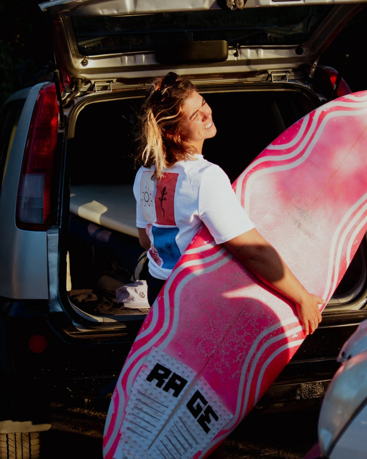 Frau mit pinkem Surfboard im Sonnenlicht trägt weißes Rebel Elements T-Shirt mit Grafik