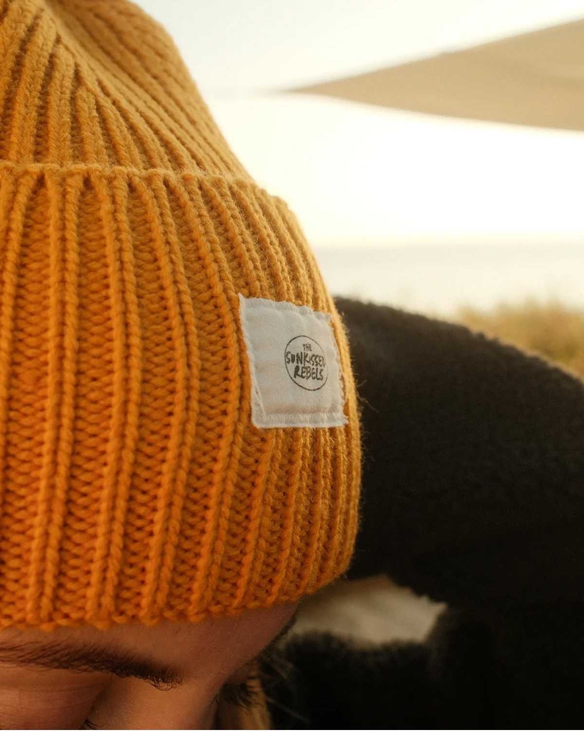 Nahaufnahme der senfgelben Rebels Big Beanie Honey Gold von The Sunkissed Rebels mit weißem Logo-Patch und Rippstrick.