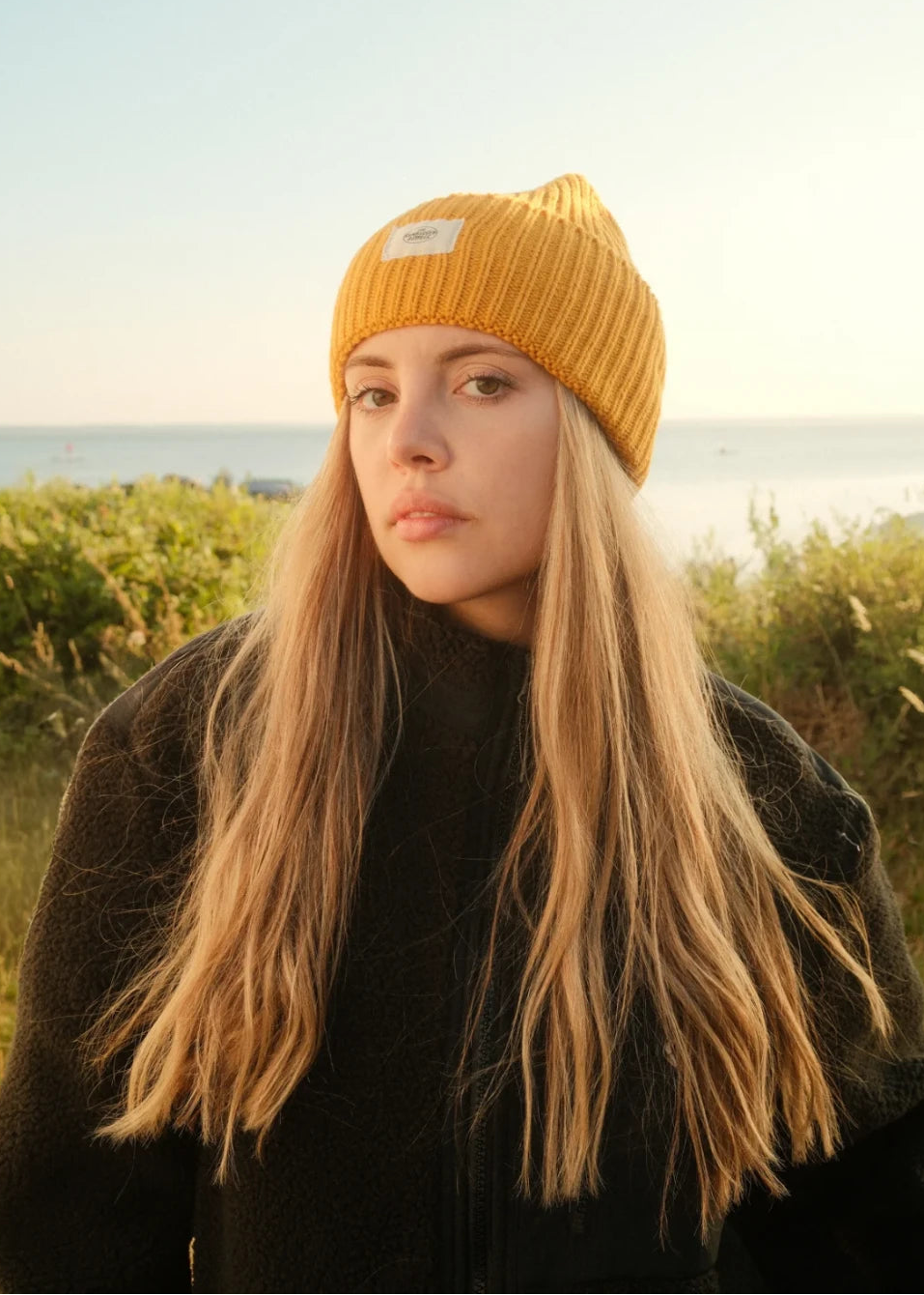 Nahaufnahme der senfgelben Rebels Big Beanie in Honey Gold von The Sunkissed Rebels mit hellem Logo-Patch und schwarzer Teddy Fleecejacke vor Meerblick.
