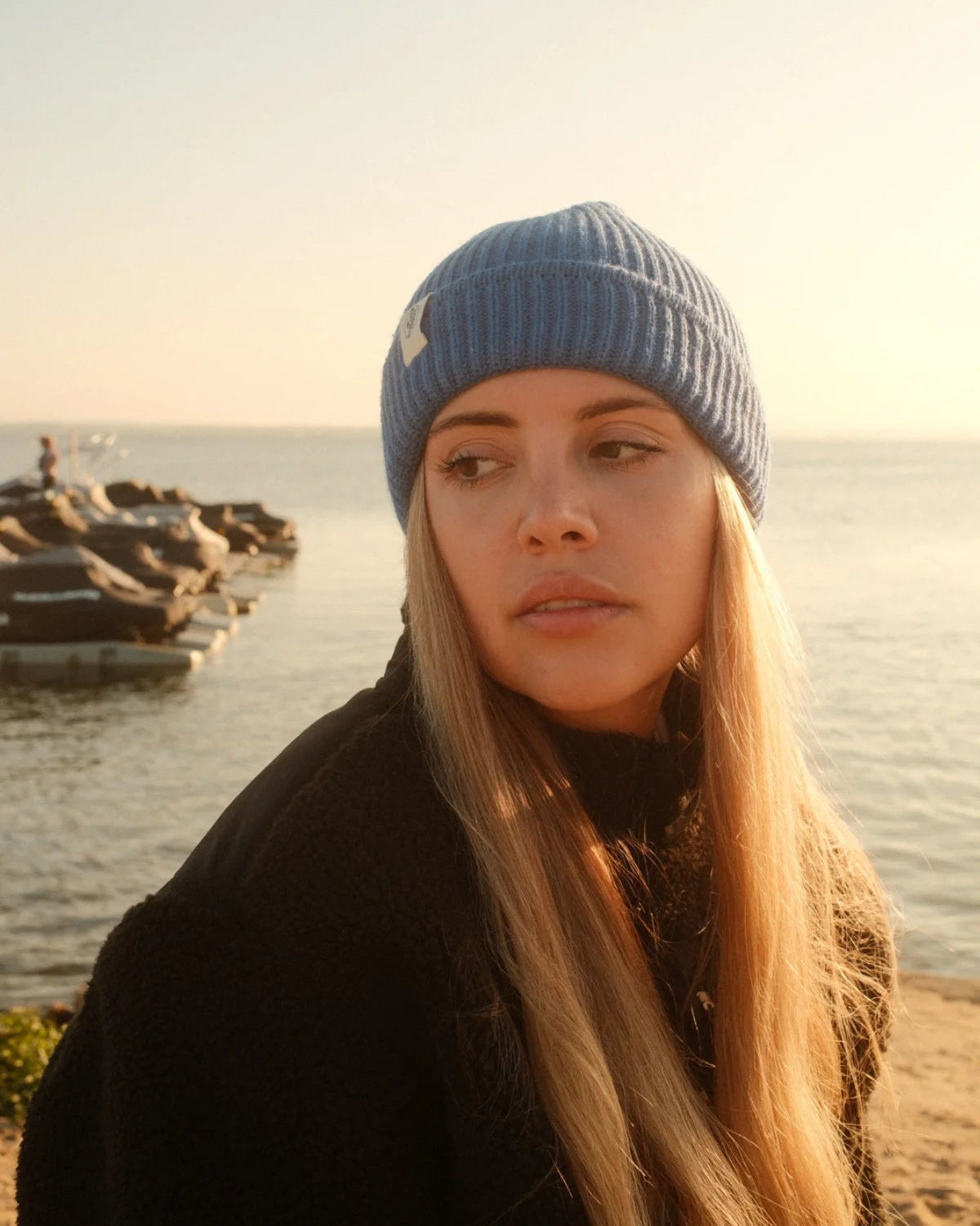 Frau am Strand mit blauer Strickmütze Rebels Knit Beanie in Ocean Blue von The Sunkissed Rebels mit Label-Patch.