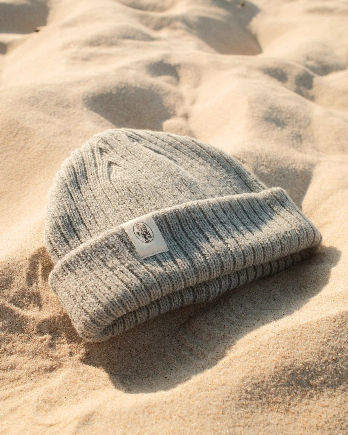 Graue Strickmütze Rebels Knit Beanie in Stormy Grey von The Sunkissed Rebels mit hellem Logo-Patch im Sand.