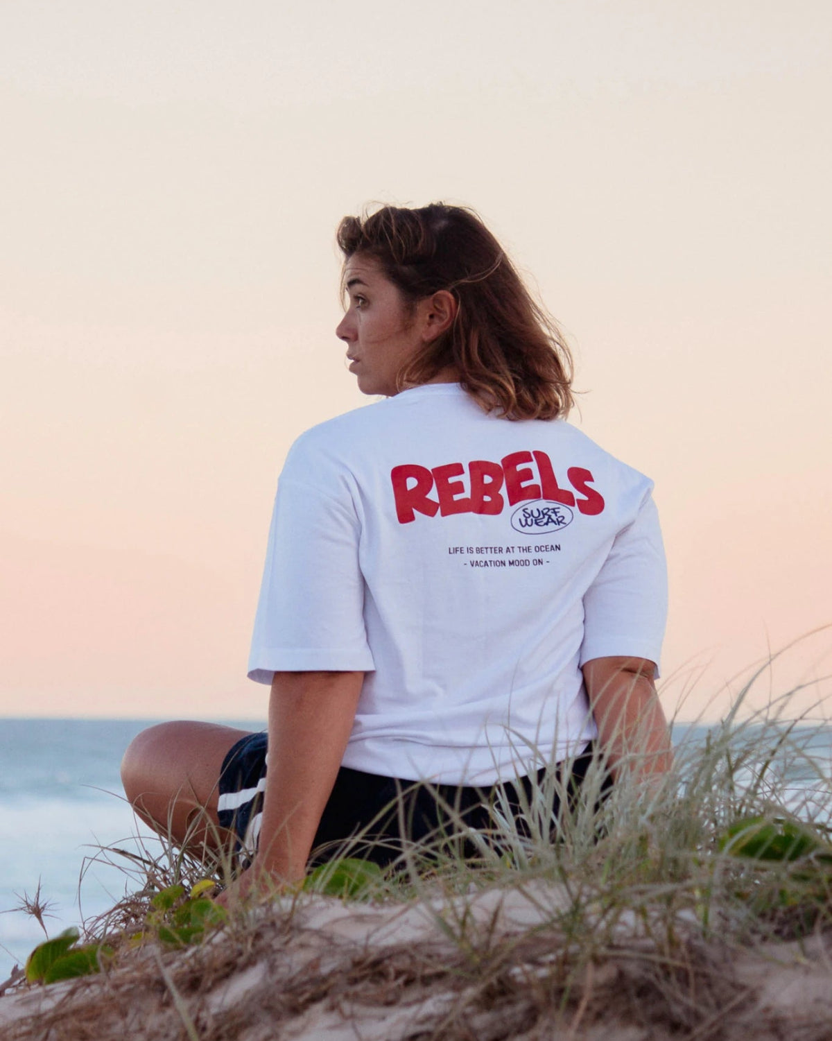 Rückansicht vom weißen Rebels Surf Wear Oversized T-Shirt mit Backprint am Meer bei Sonnenuntergang