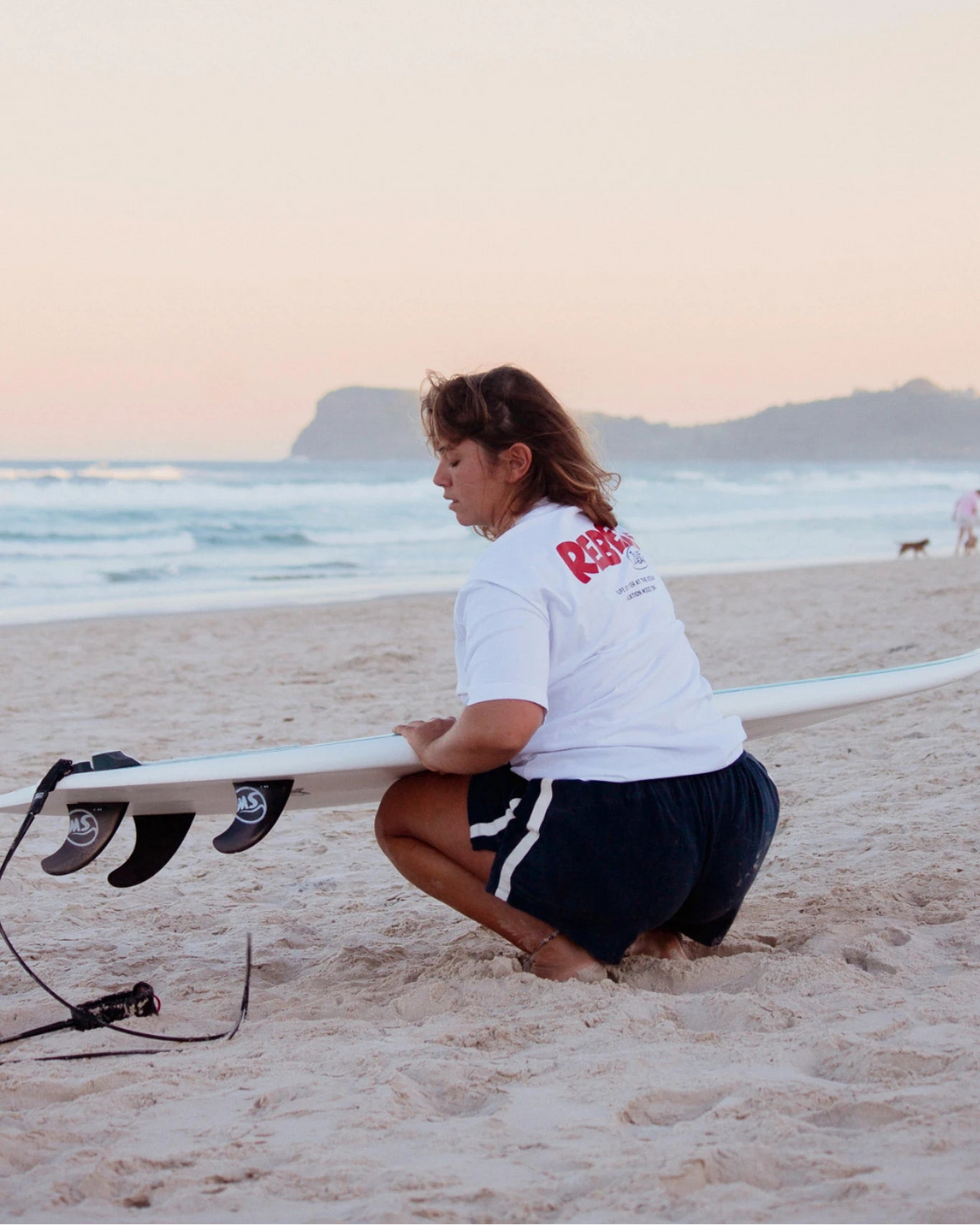 Frau am Strand mit Surfboard trägt weißes Rebels Surf Wear Oversized T-Shirt im Sonnenuntergang
