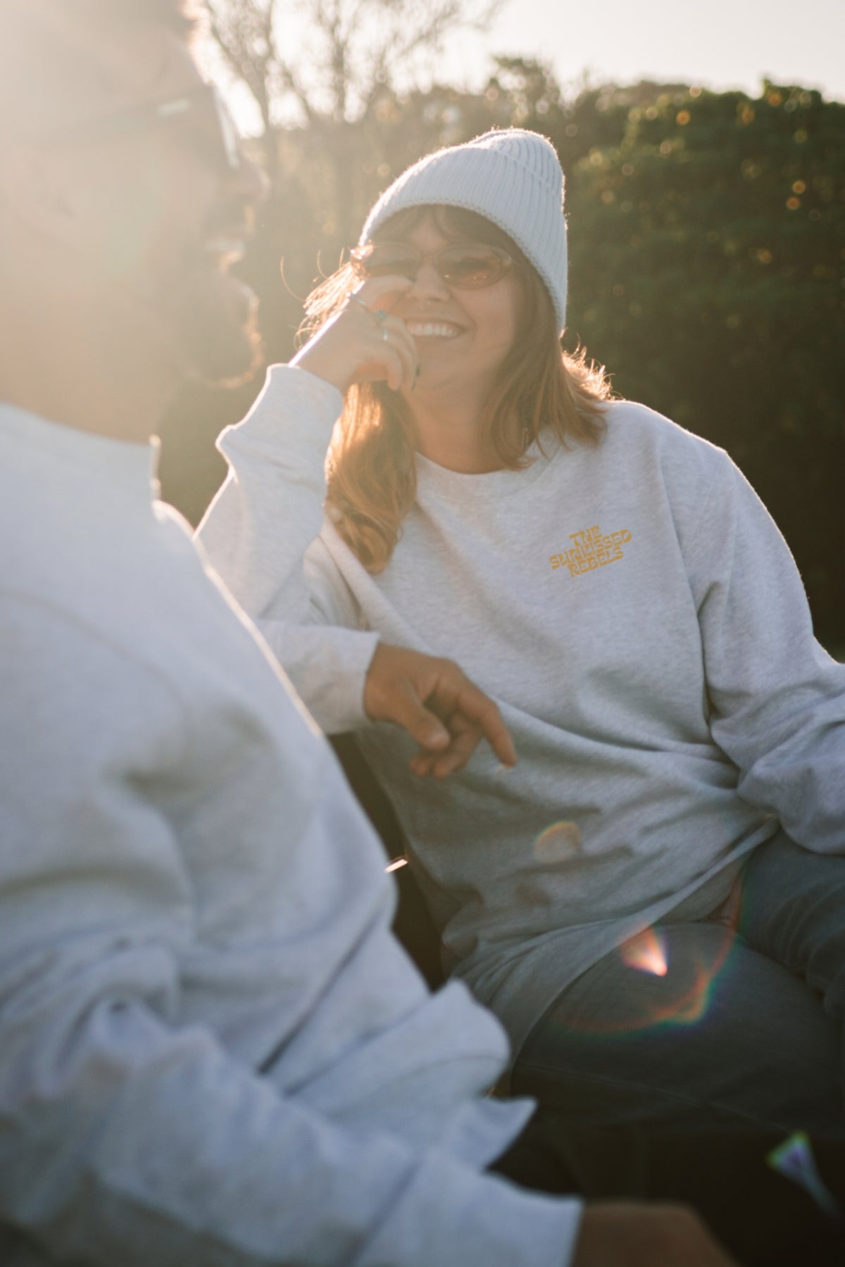 Frau sitzt mit Freund auf einer Bank. Beide tragen Sweater von The Sunkissed Rebels aus der Paradise & Sunset Kollektion.