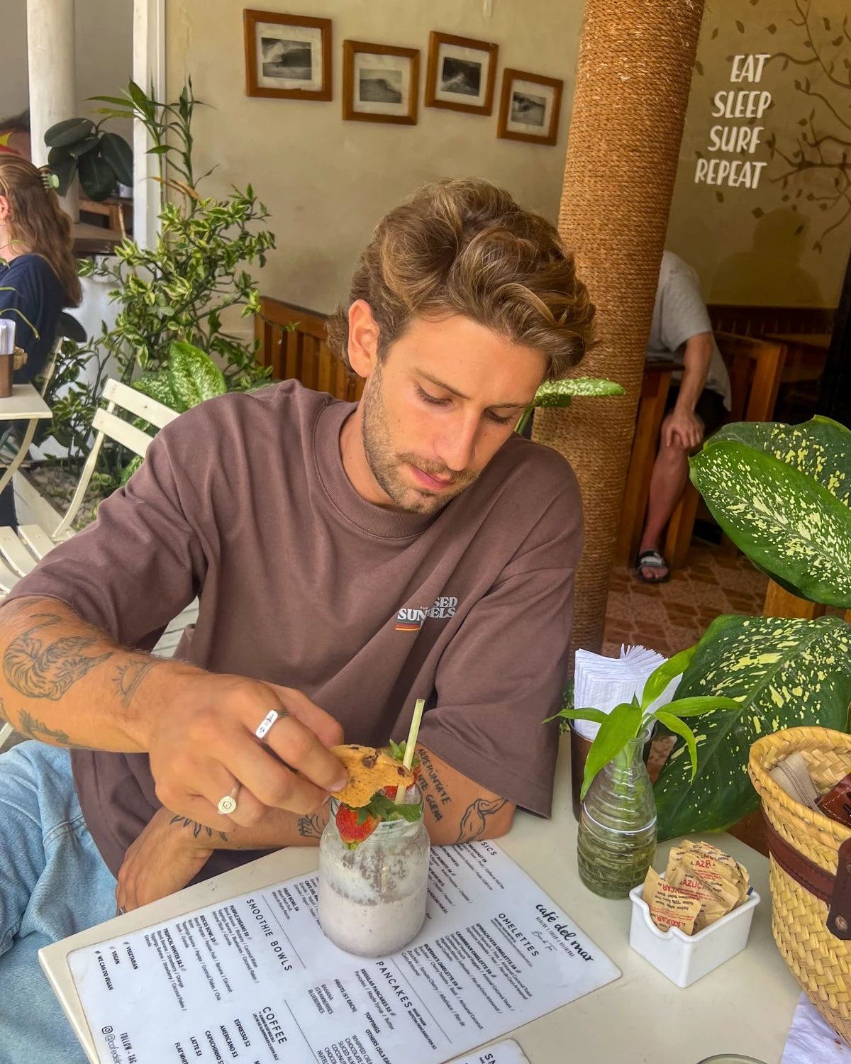 Mann trägt das Retro Vibes Heavy T-Shirt in Mocha beim Frühstück in einem tropischen Café mit Smoothie und Cookie.