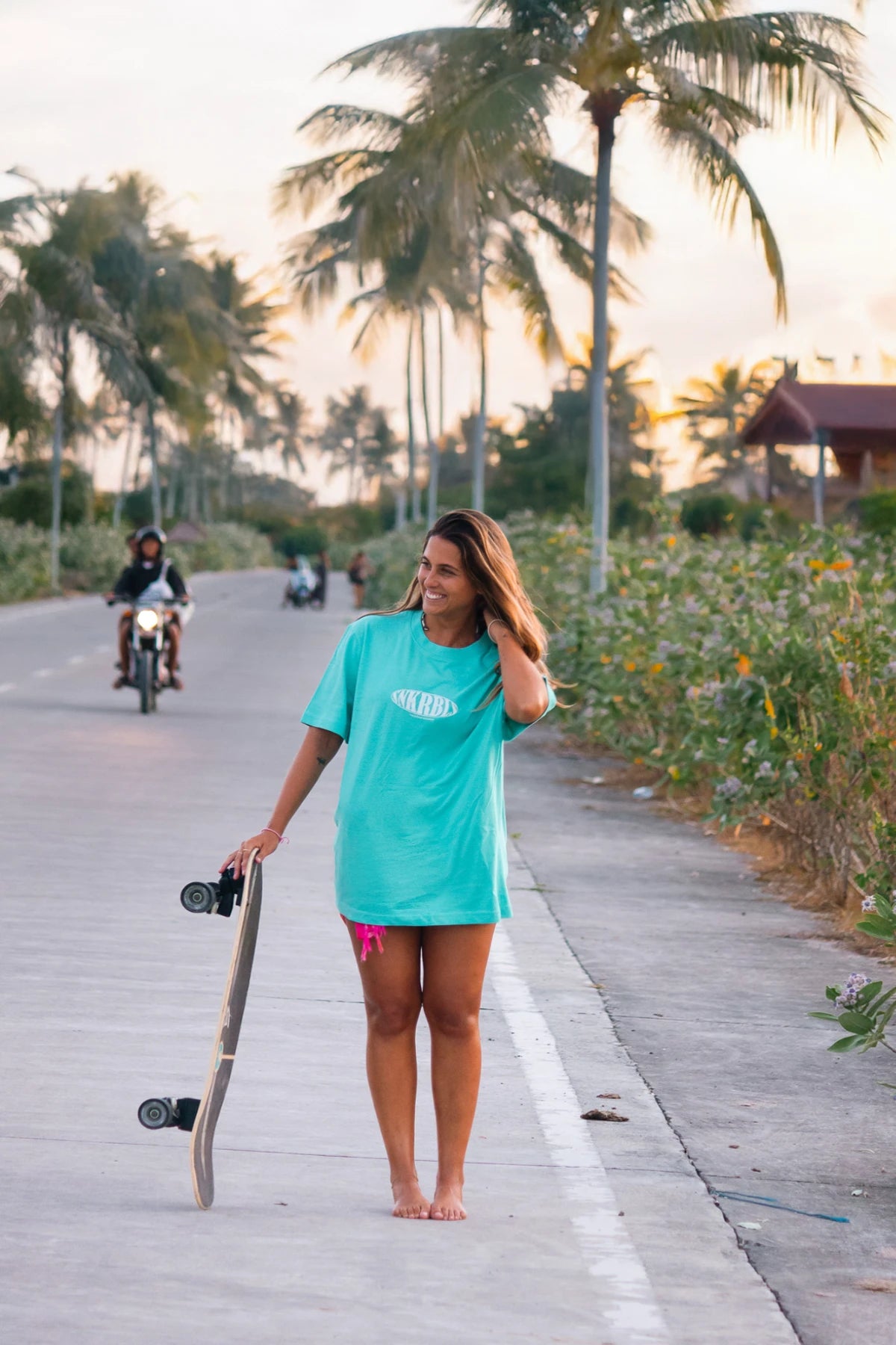 Frau im türkisfarbenen SNKRBLs T-Shirt mit Skateboard bei Sonnenuntergang