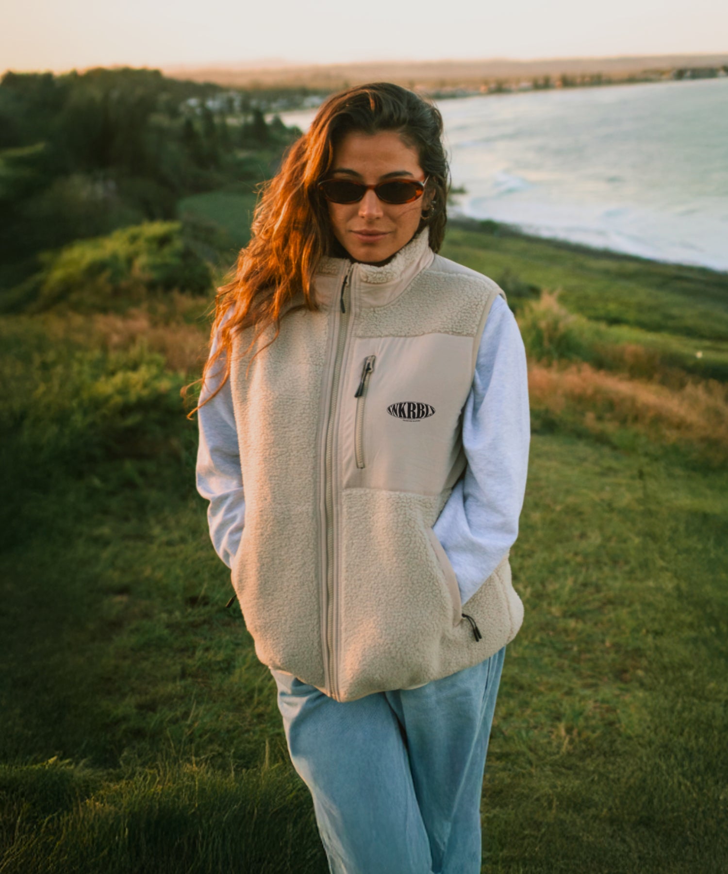 Frau trägt eine beige Sherpa Fleece Vest von The Sunkissed Rebels mit Blick auf die Küste während des Sonnenuntergangs.