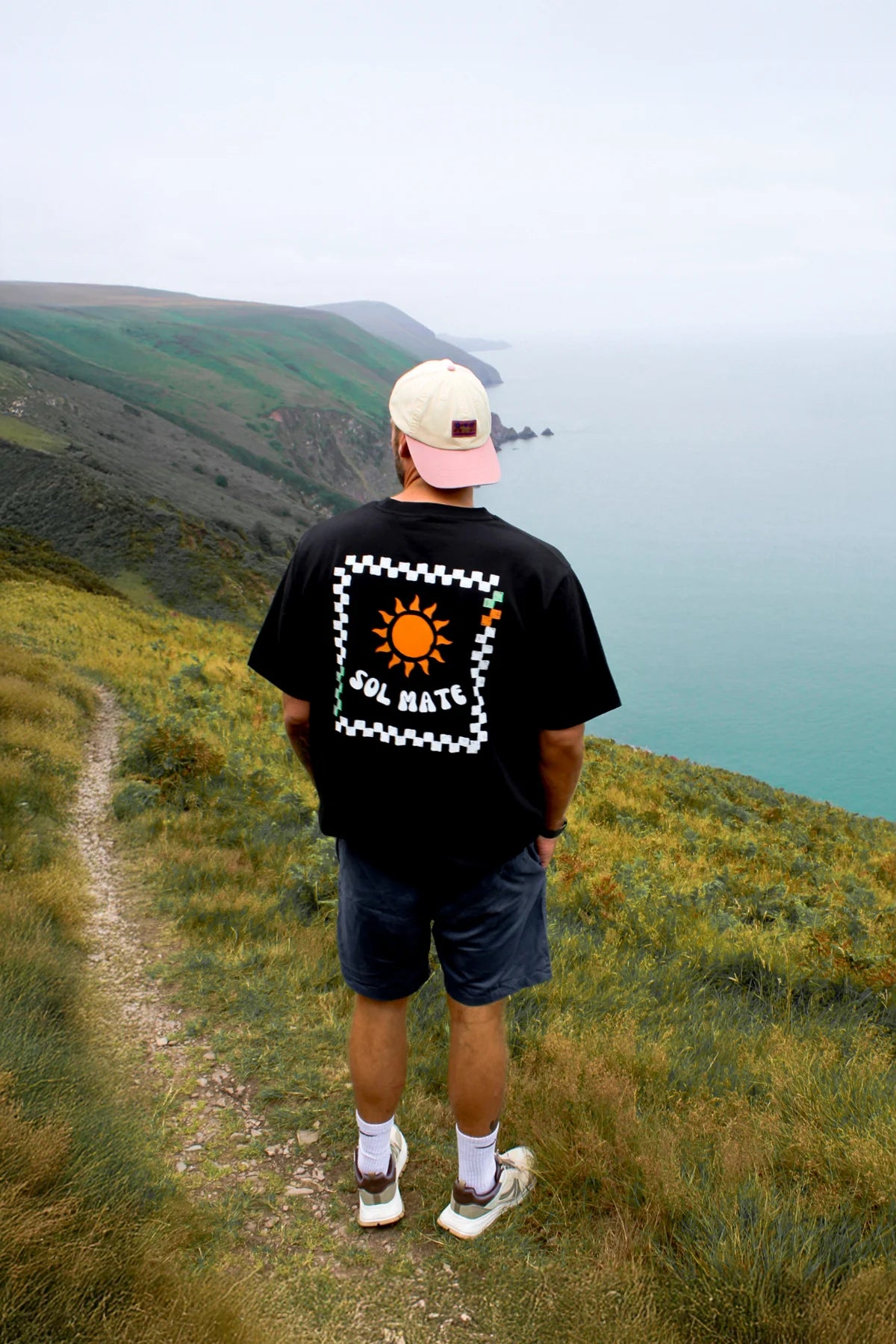 Rückansicht des Sol Mate Oversized T-Shirts in Schwarz mit großem Sonnenmotiv, Mann blickt aufs Meer von einer Klippe.