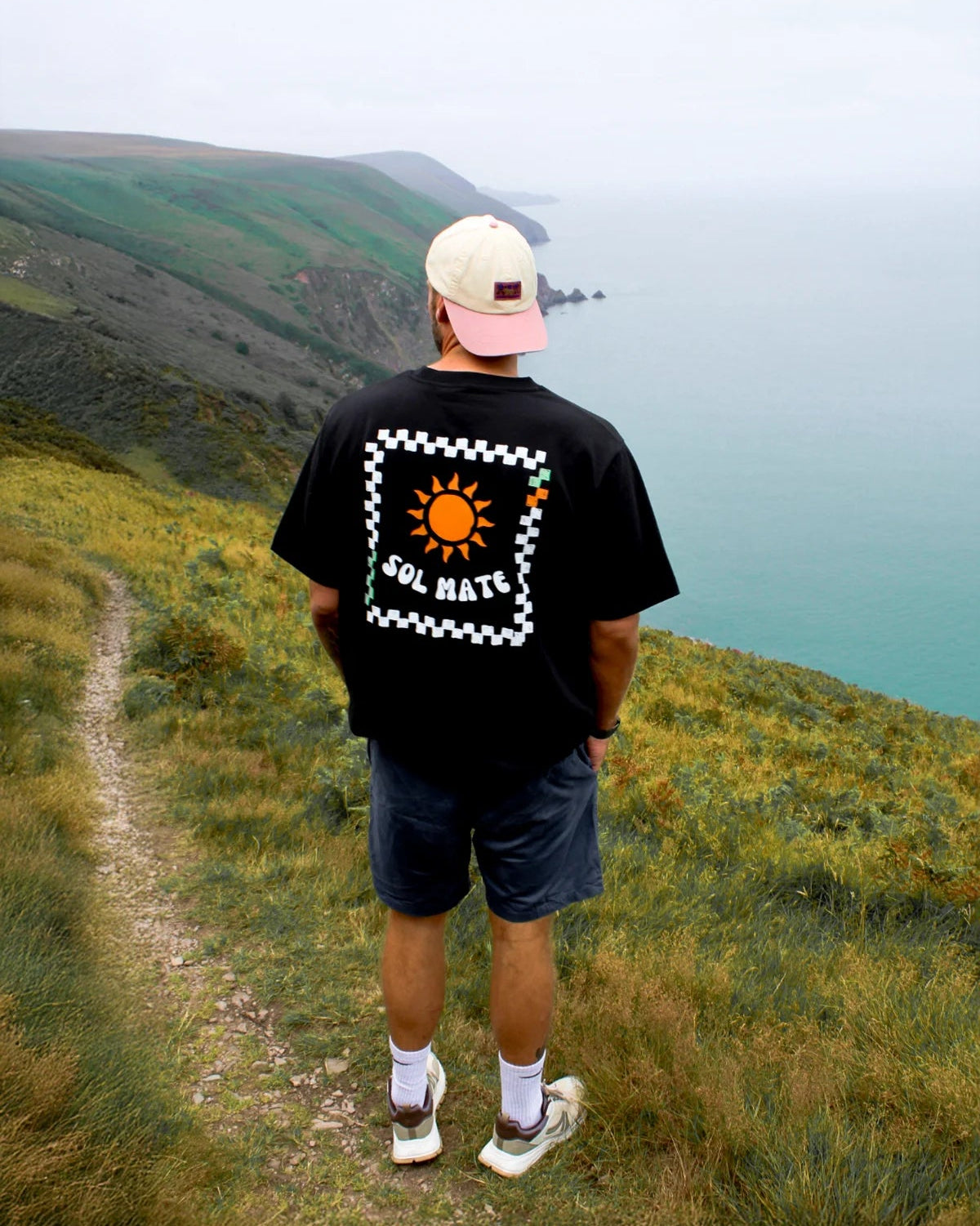 Rückansicht des Sol Mate Oversized T-Shirts in Schwarz mit großem Sonnenmotiv, Mann blickt aufs Meer von einer Klippe.