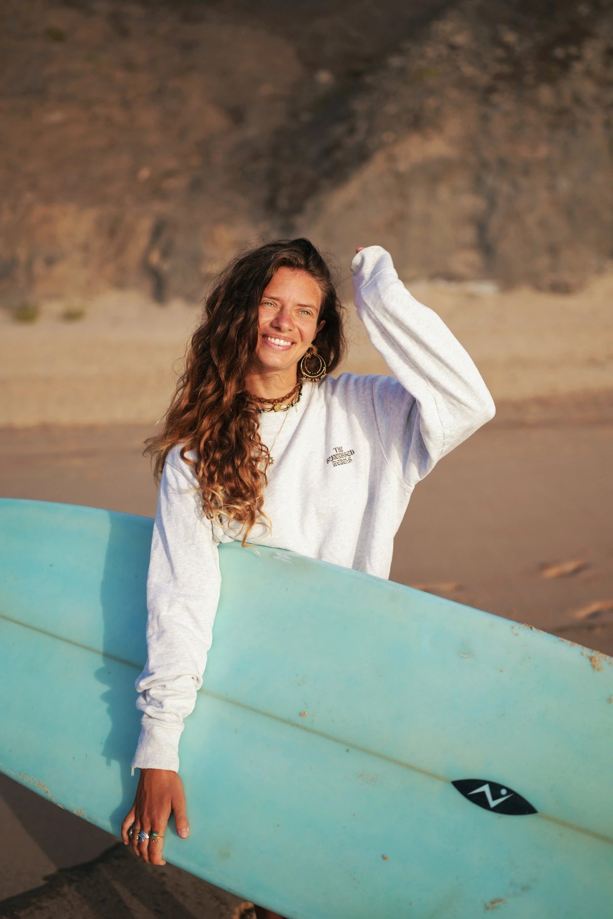 Frau mit offenen Haaren und Surfbrett im Paradise & Sunset Pullover