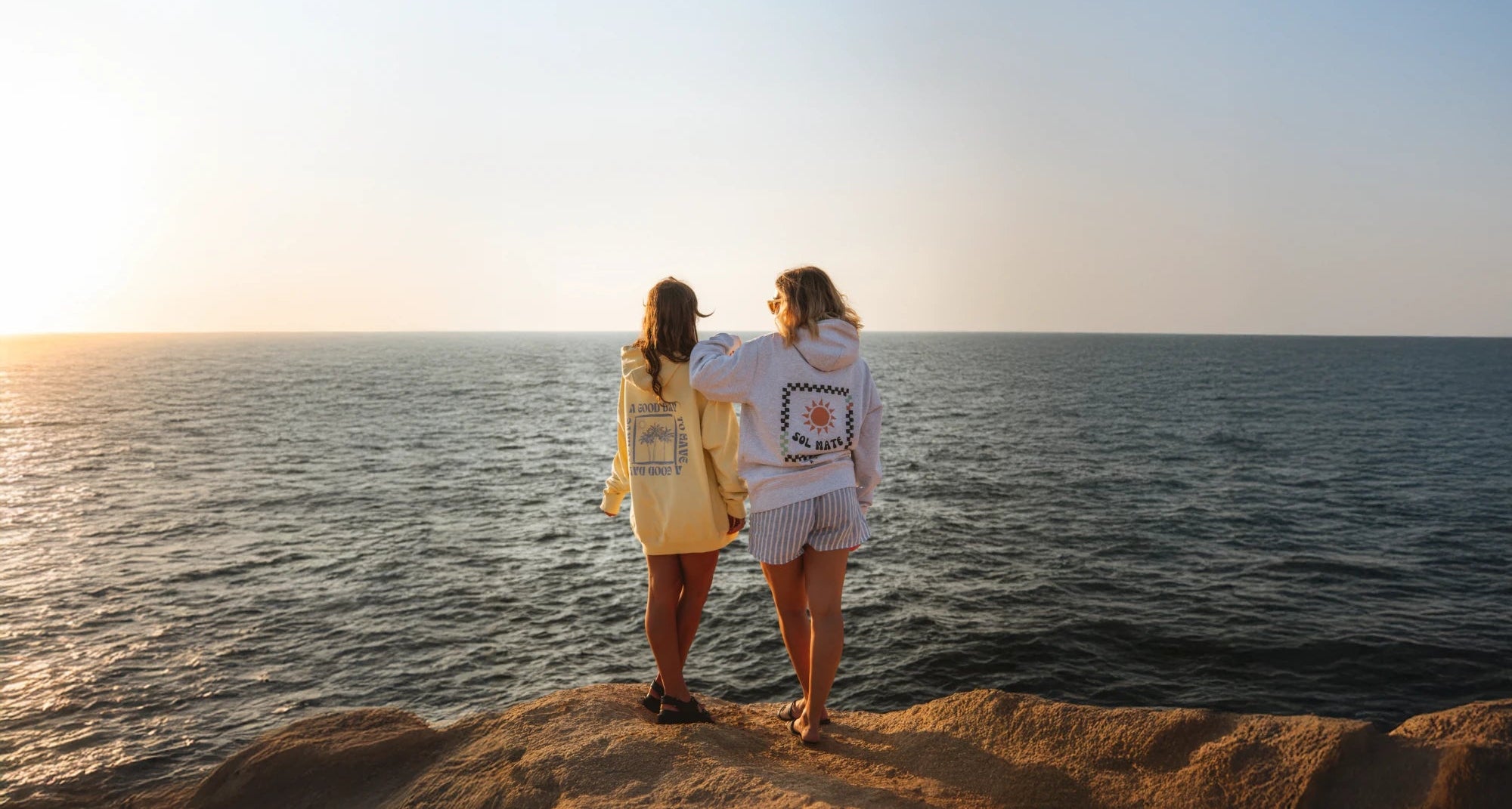 Zwei Models von The Sunkissed Rebels stehen bei Sonnenuntergang auf einer Klippe am Meer und tragen Hoodies der aktuellen Kollektion, mit Blick auf den Ozean.