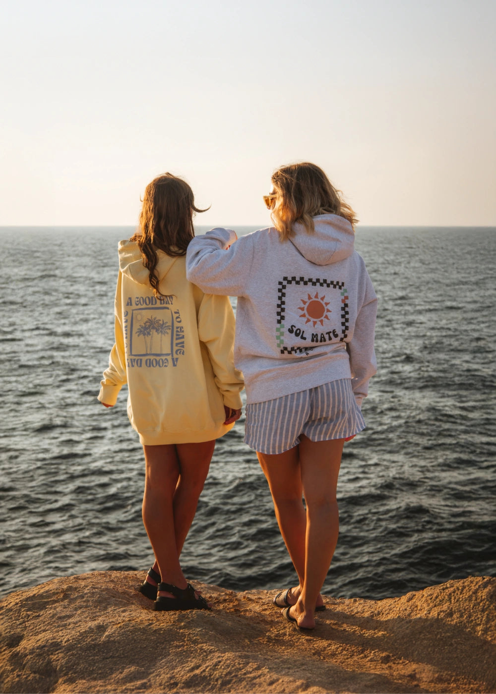 Zwei Models von The Sunkissed Rebels stehen auf einer Klippe mit Blick aufs Meer und tragen sommerliche Hoodies der Kollektion, eingefangen im warmen Licht des Sonnenuntergangs.