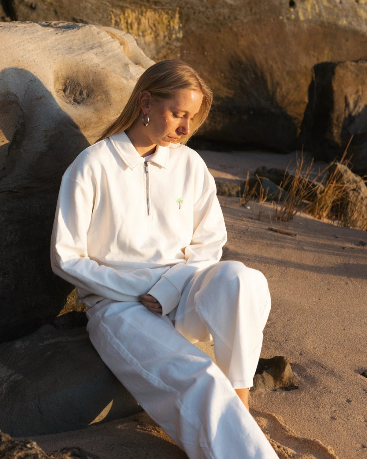 Frau sitzt am Strand an Felsen und trägt cremefarbenen Half-Zip Pullover im warmen Abendlicht
