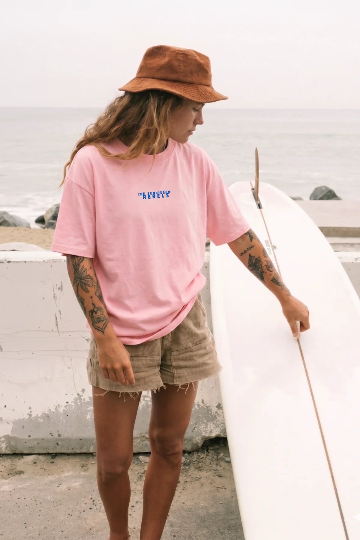 Frau mitz Surfbrett trägt pinkes Oversized T-Shirt von The Sunkissed Rebels