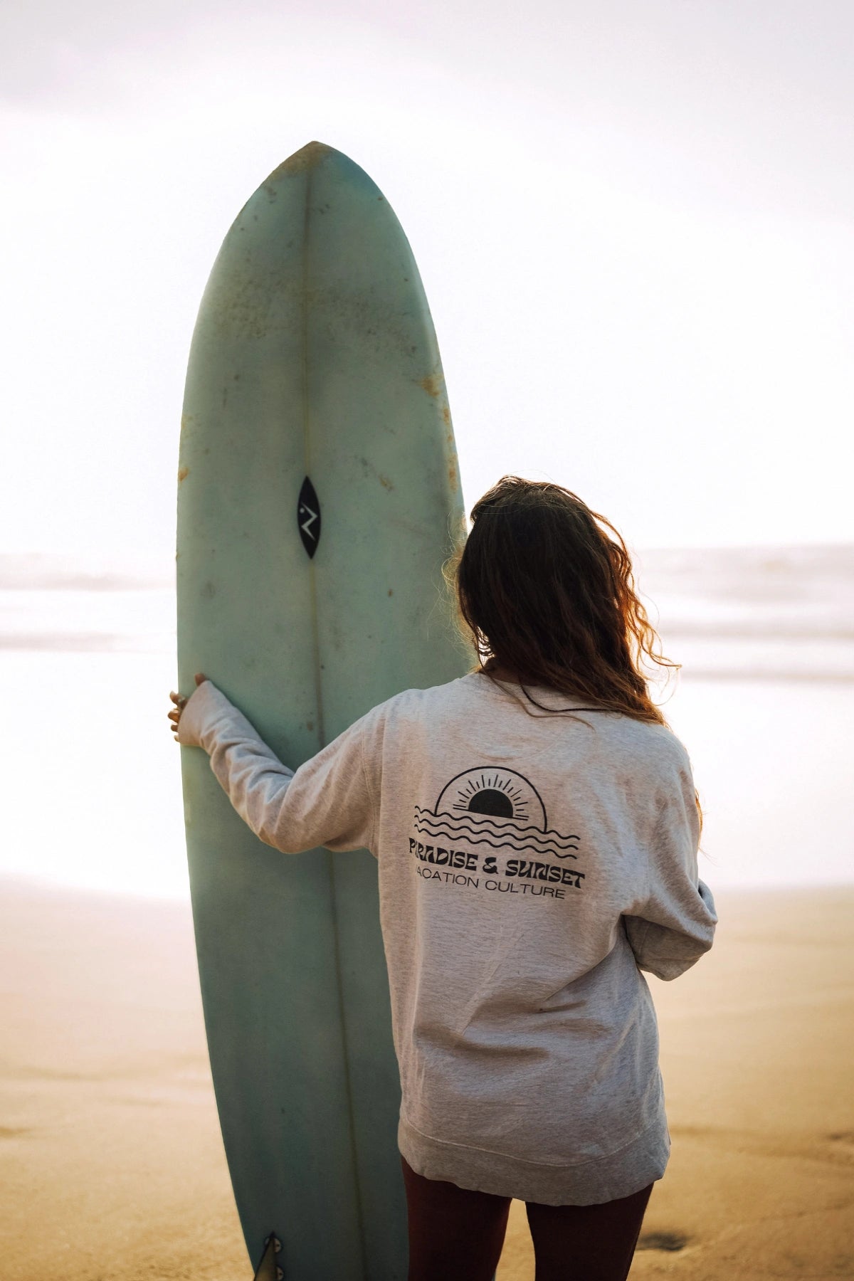 Rückenansicht einer Frau mit Surfbrett im Paradise & Sunset Sweater bei Sonnenaufgang