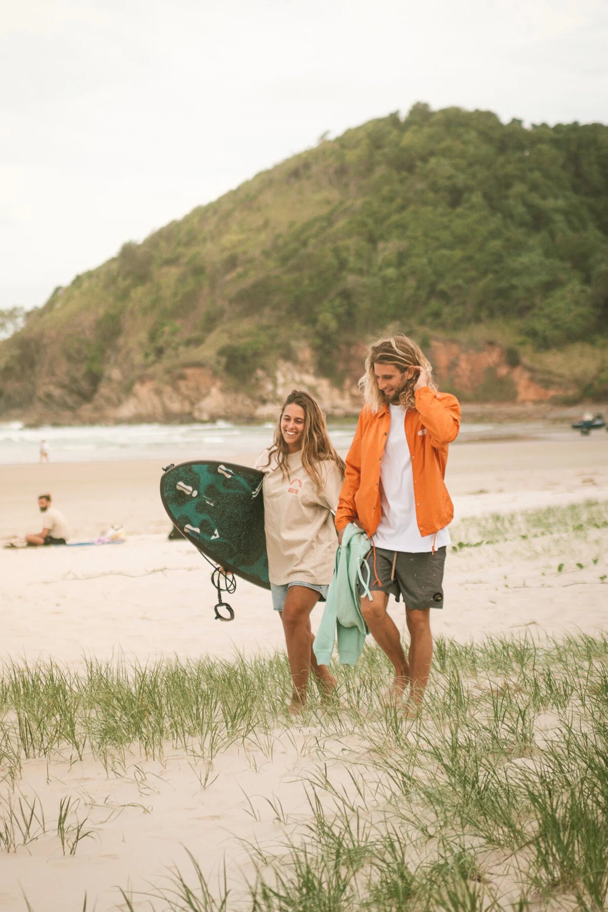 Pärchen mit The Sunkissed Rebels T-Shirts am Strand mit Surfbrett. Mann trägt orangene Coach Jacket aus recyceltem Material.