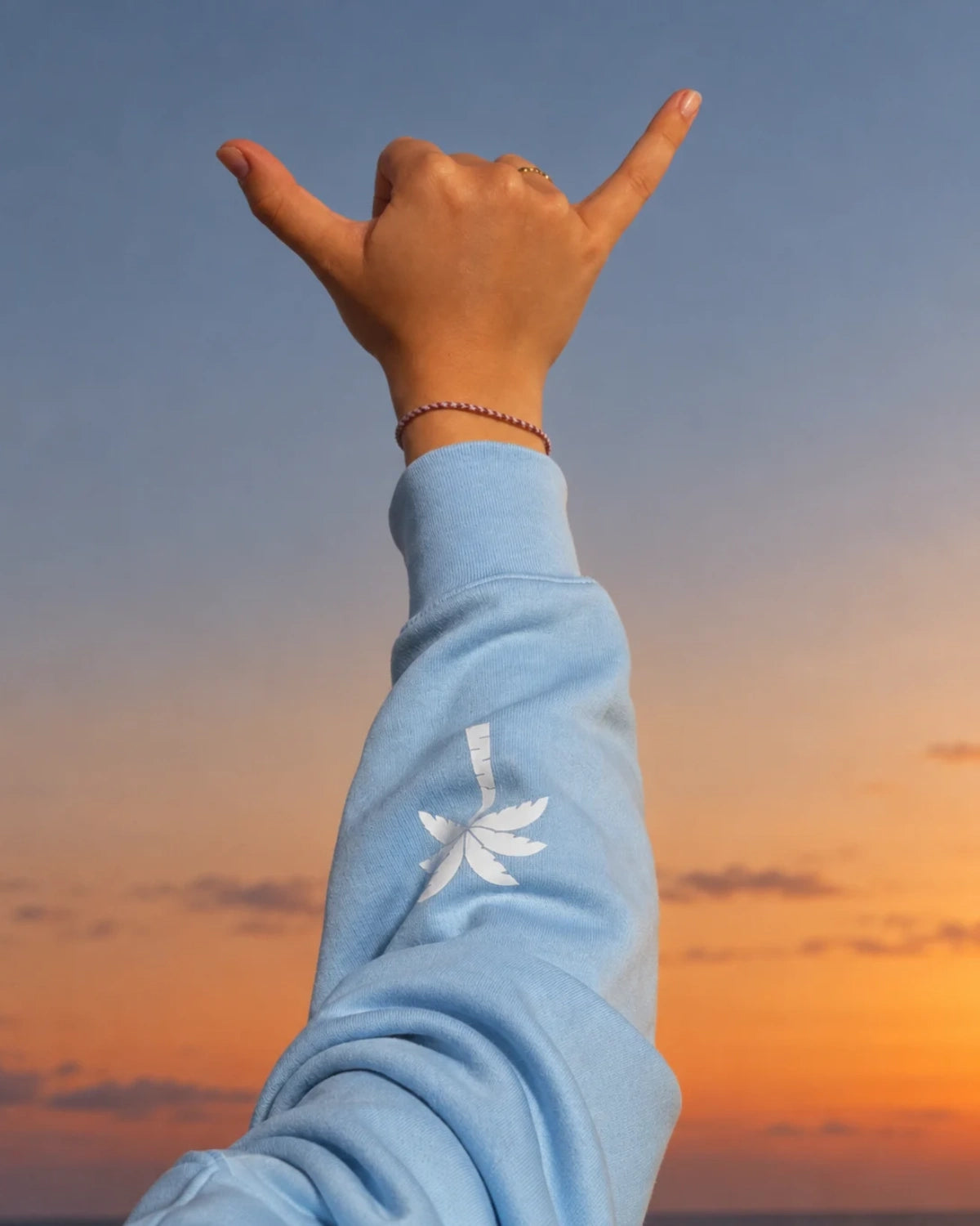 Detailaufnahme des The Horizon Hoodie – Blue Soul mit weißem Palmen-Print am Ärmel vor warmem Abendhimmel.
