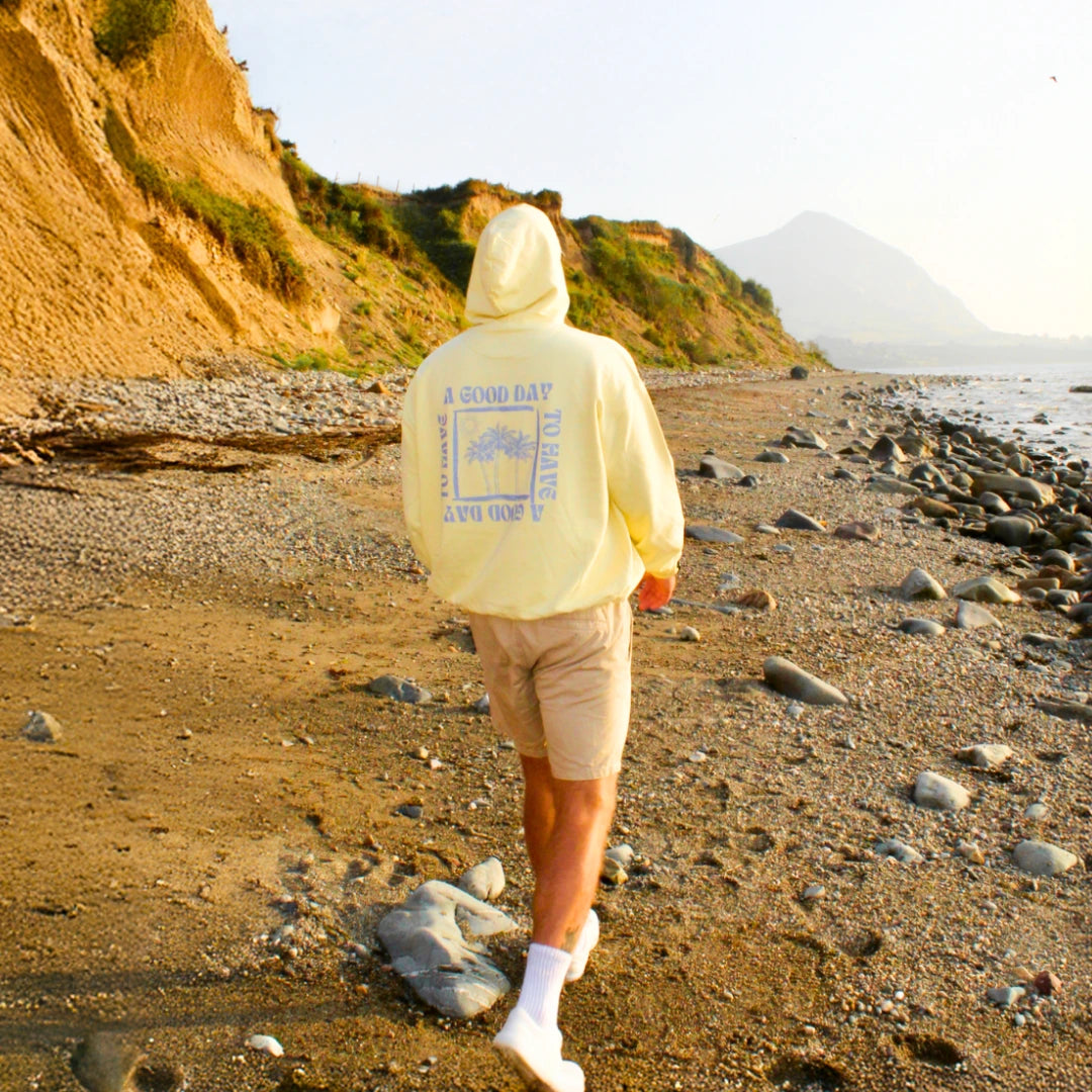 Person läuft am Strand im gelben The Sunkissed Rebels Hoodie mit großem Rückenprint entlang der Küste.