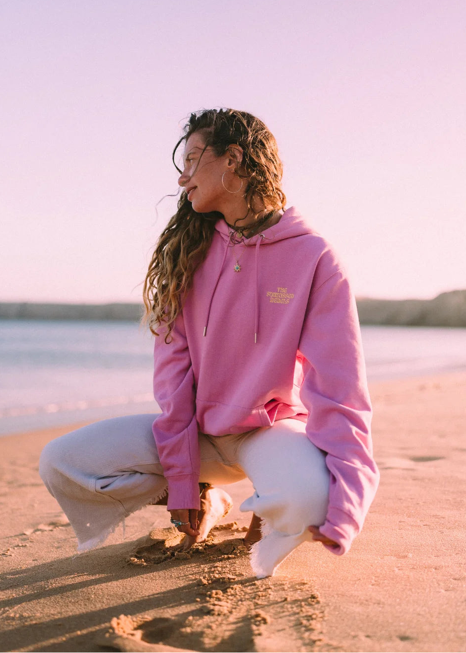 Frau trägt den pinken Oversized Hoodie von The Sunkissed Rebels an einem Sandstrand während des Sonnenuntergangs im Beach-Lifestyle.