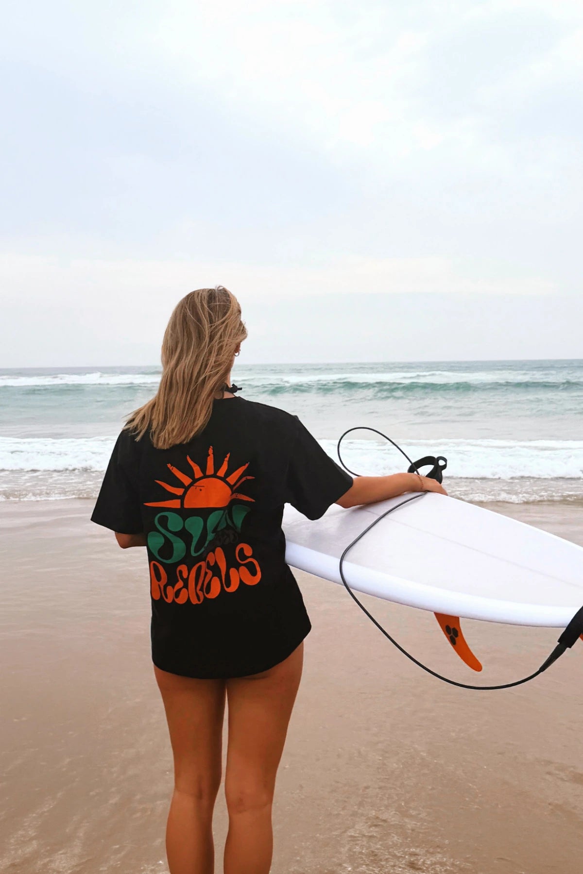Frau trägt das The Sunkissed Rebels Oversized T-Shirt Black mit großem farbigem Rückenprint und Surfboard am Strand vor dem Meer.