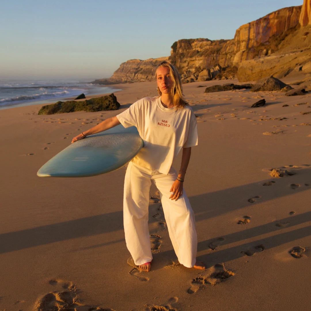Frau mit Surfboard am goldenen Sandstrand bei Sonnenuntergang im weißen The Sunkissed Rebels T-Shirt.