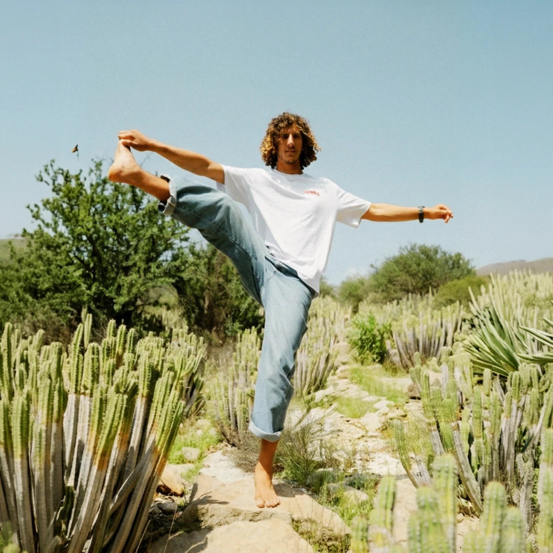 Mann balanciert barfuß in einer Yoga-Pose zwischen Kakteen und trägt ein weißes The Sunkissed Rebels T-Shirt.