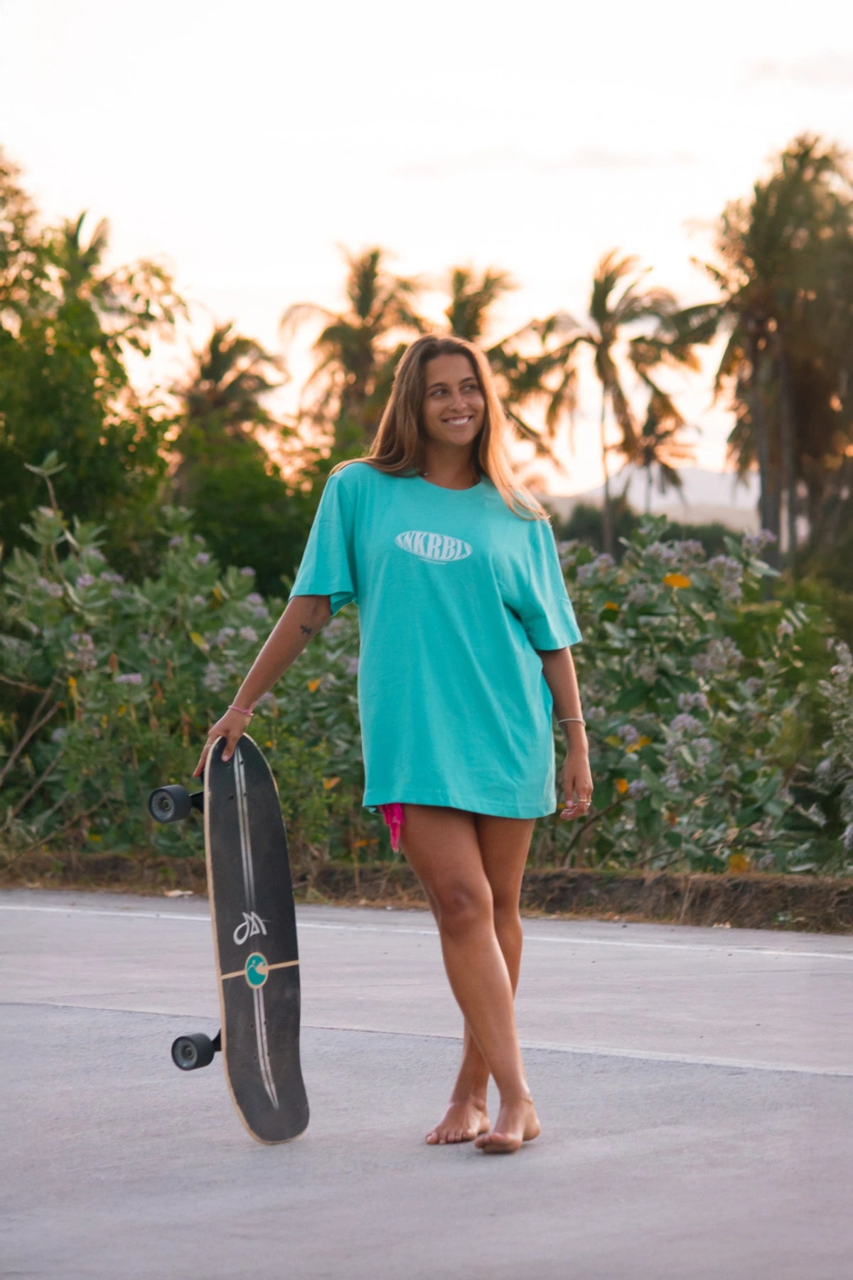 Frau barfuß mit Skateboard im SNKRBLs Pool Blue Shirt auf Palmenstraße