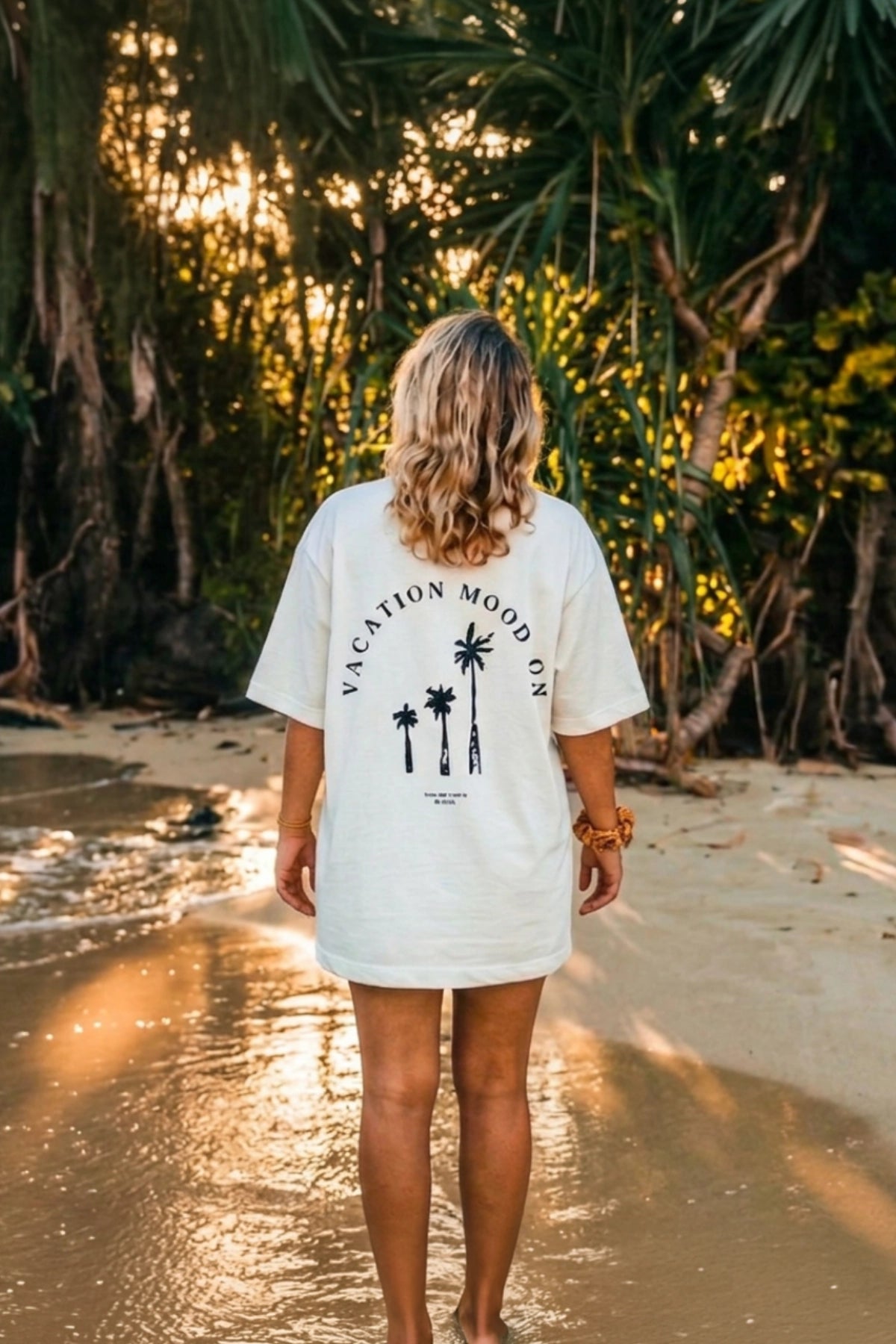 Frau trägt das Vacation Mood On Boxy Oversized T-Shirt in White von The Sunkissed Rebels mit Palmen-Backprint an einem tropischen Strand.