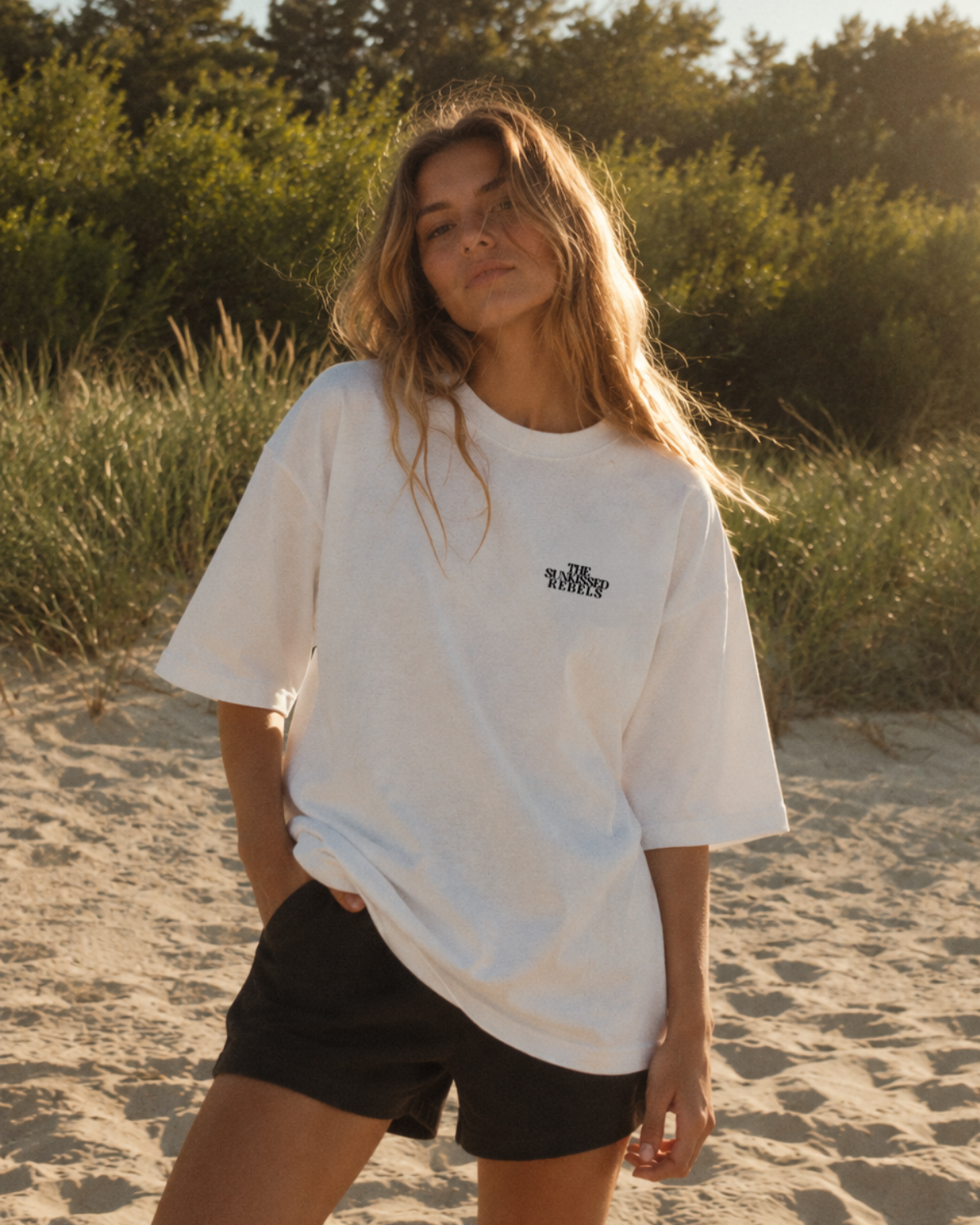 Frau am Strand im weißen Vacation Mood On Boxy Oversized T-Shirt von The Sunkissed Rebels mit schwarzem Brustlogo und weiten Ärmeln.