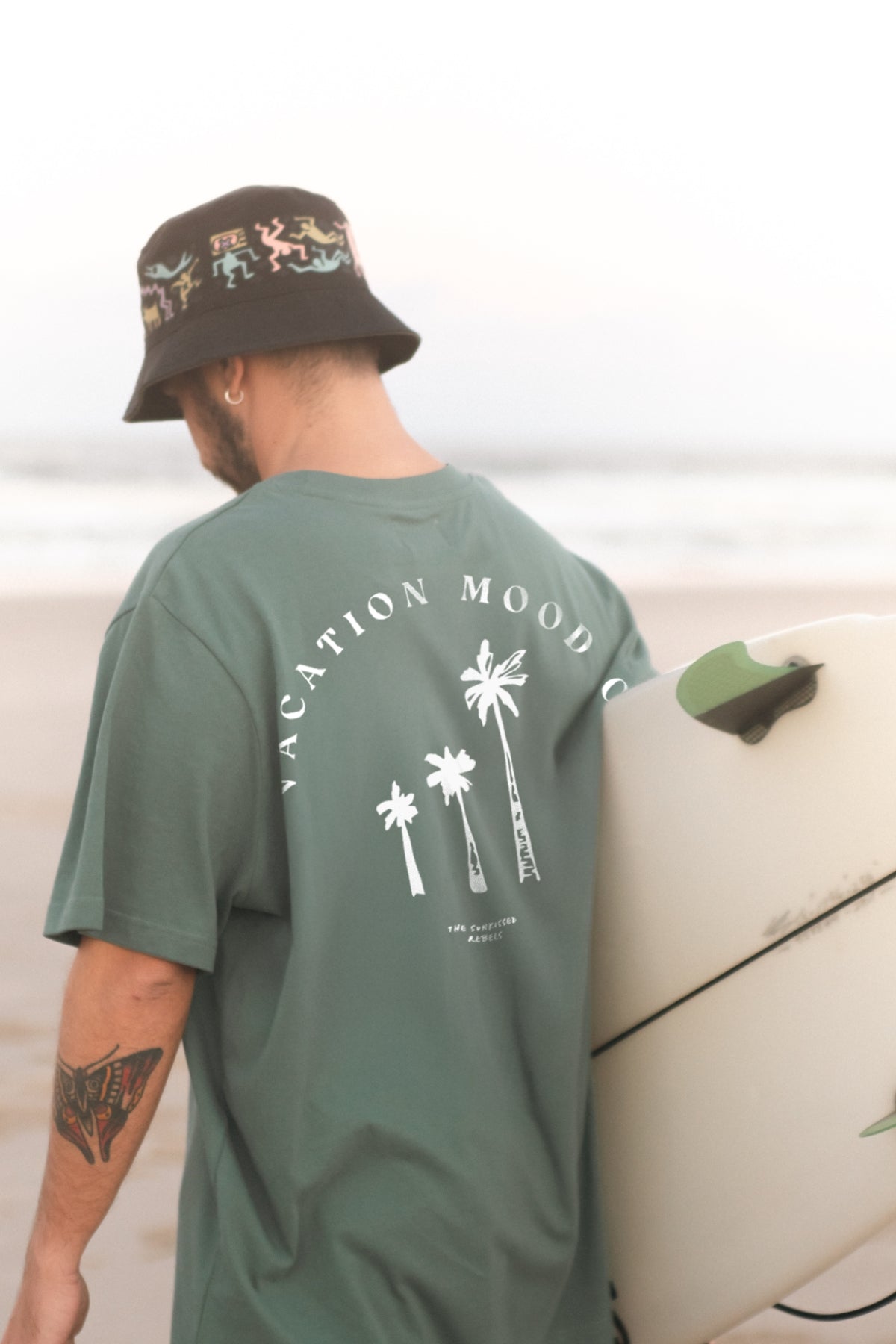 Mann hält ein Surfbrett unter dem Arm und trägt das Vacation Mood On T-Shirt von The Sunkissed Rebels