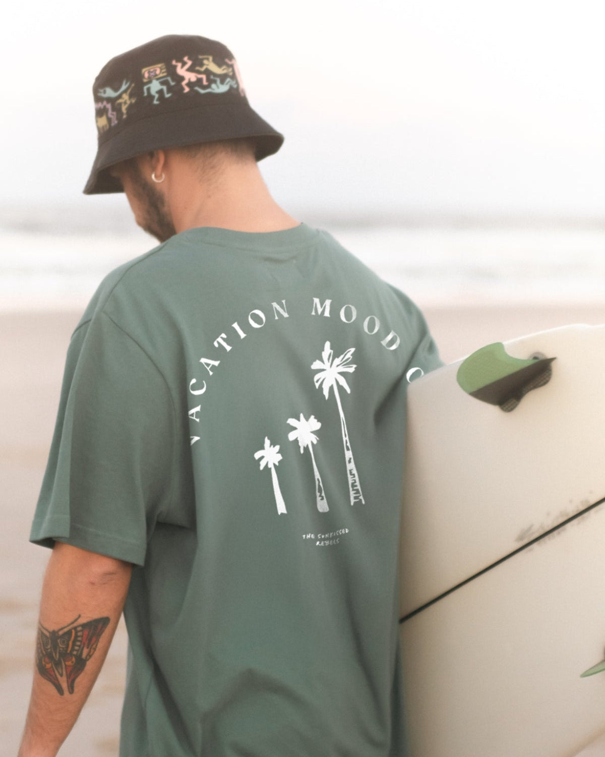 Mann hält ein Surfbrett unter dem Arm und trägt das Vacation Mood On T-Shirt von The Sunkissed Rebels