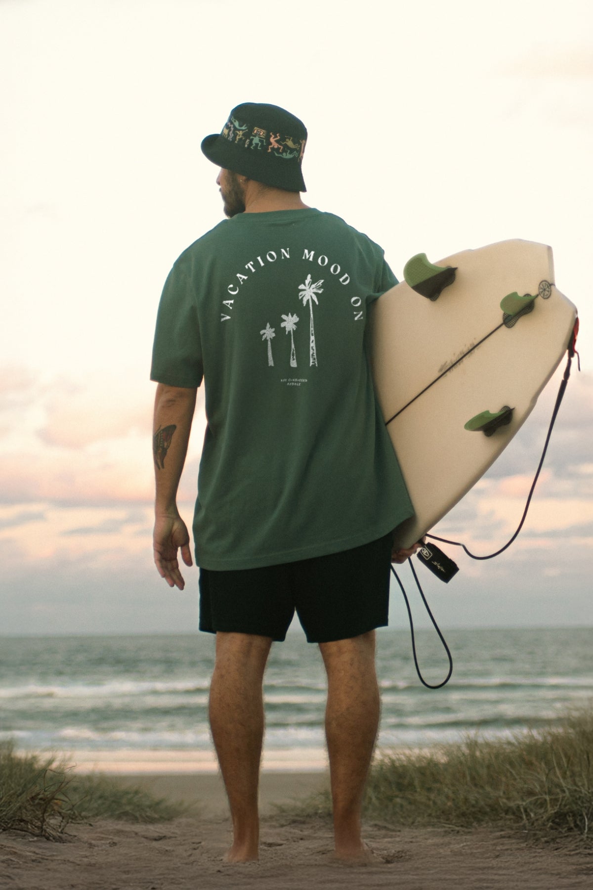 Mann trägt Vacation Mood On T-Shirt in Green Bay von The Sunkissed Rebels am strand und hält ein Surfbrett unter dem Arm.