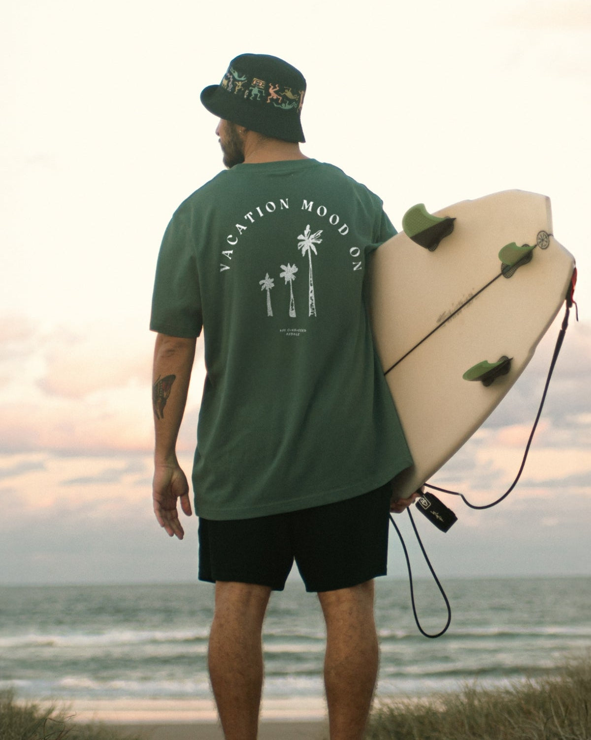 Mann trägt Vacation Mood On T-Shirt in Green Bay von The Sunkissed Rebels am strand und hält ein Surfbrett unter dem Arm.