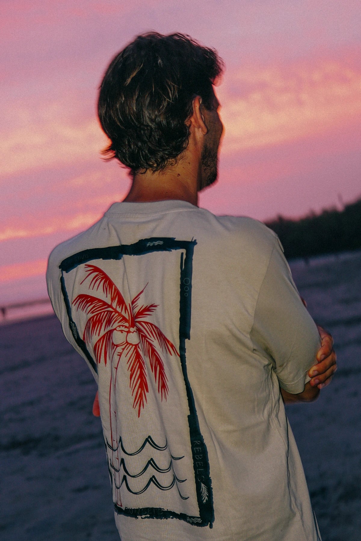 Mann trägt das Waves & Palm Trees Oversized T-Shirt in Sand mit großem Rückenprint am Strand bei Sonnenuntergang.