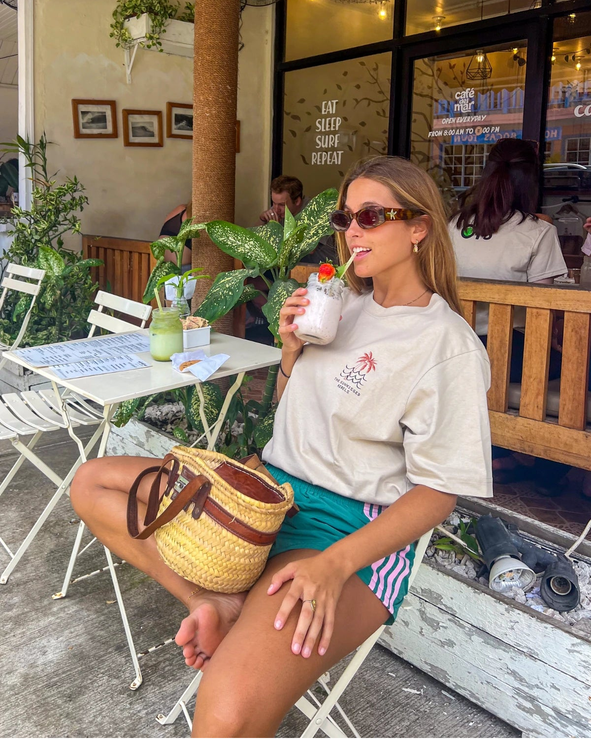 Frau trägt das Waves & Palm Trees Oversized T-Shirt in Sand beim Frühstück in einem tropischen Café mit Smoothie und Cookie.