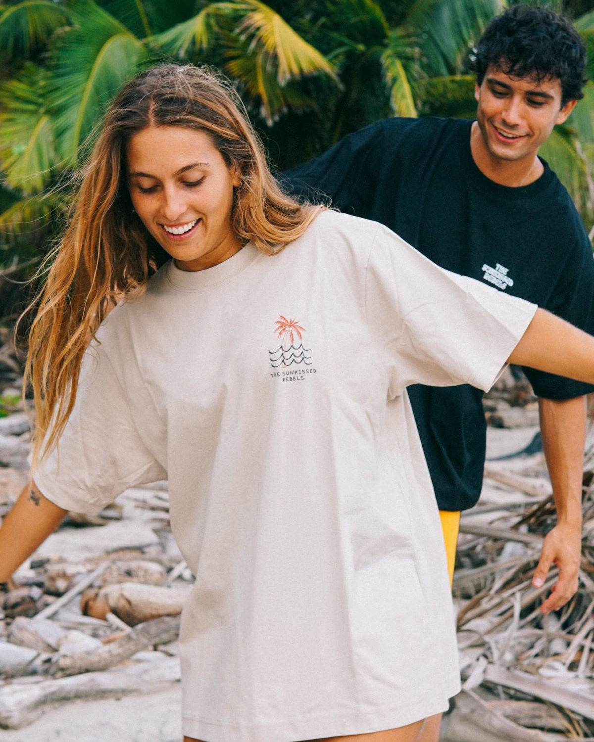 Detailaufnahme des Waves & Palm Trees Oversized T-Shirts in Sand mit Brustprint vor tropischer Strandkulisse.