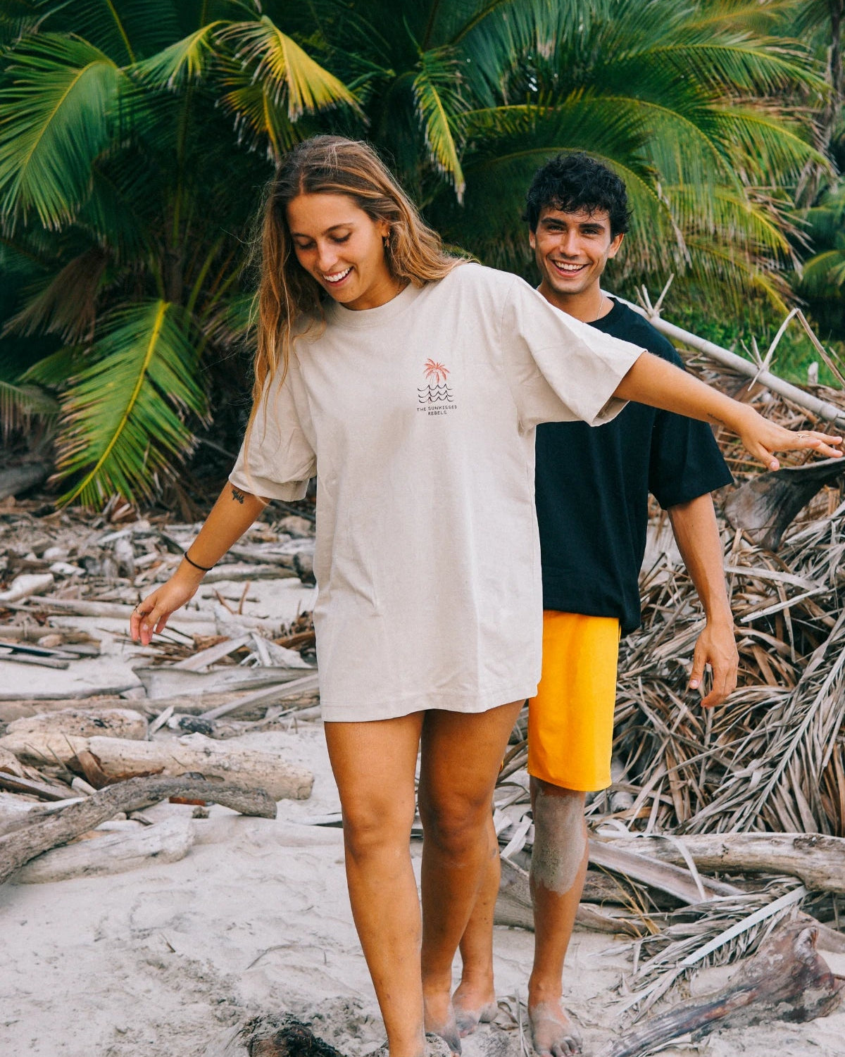 Lifestyle-Aufnahme des Waves & Palm Trees Oversized T-Shirts in Sand an einem tropischen Strand mit Palmen und Treibholz