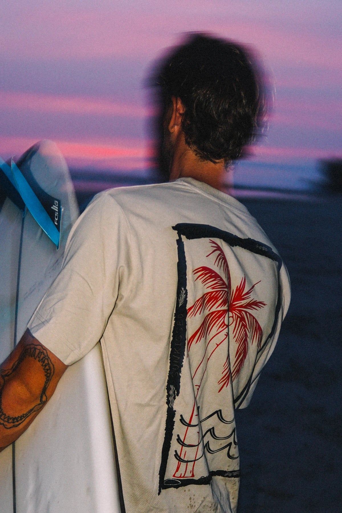 Mann trägt das Waves & Palm Trees Oversized T-Shirt in Sand mit großem Rückenprint und Surfboard bei Sonnenuntergang am Strand.
