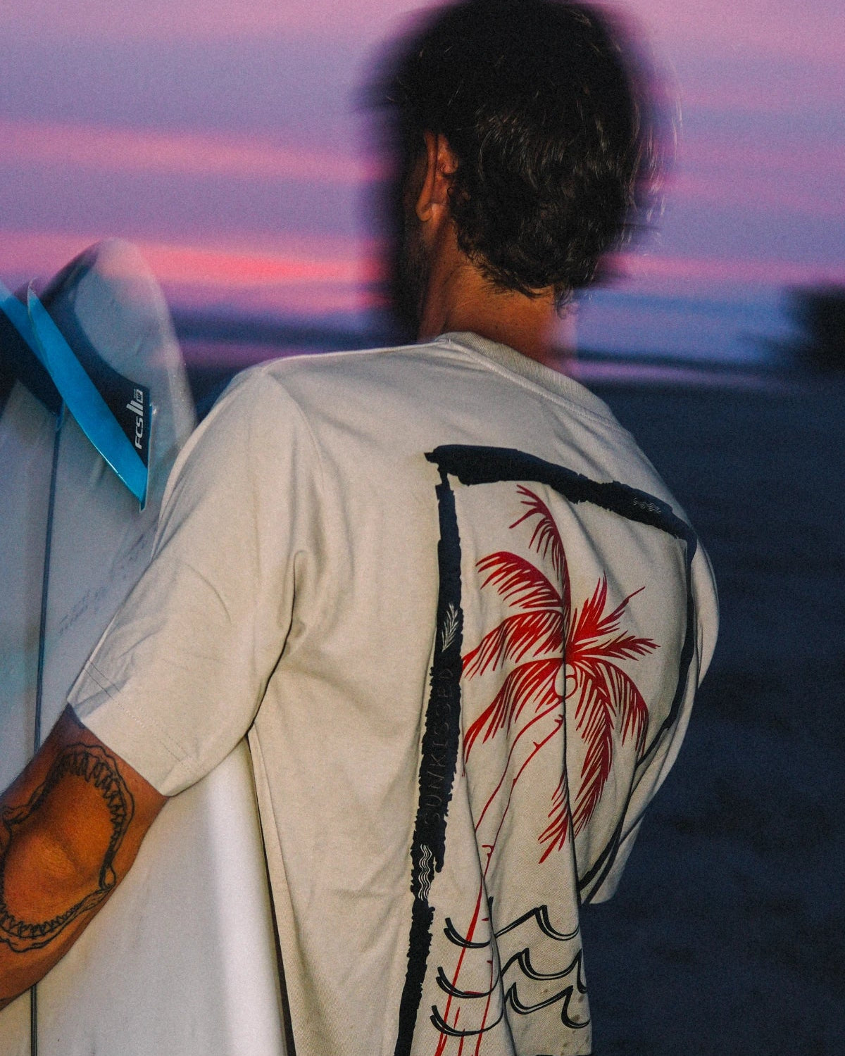 Mann trägt das Waves & Palm Trees Oversized T-Shirt in Sand mit großem Rückenprint und Surfboard bei Sonnenuntergang am Strand.