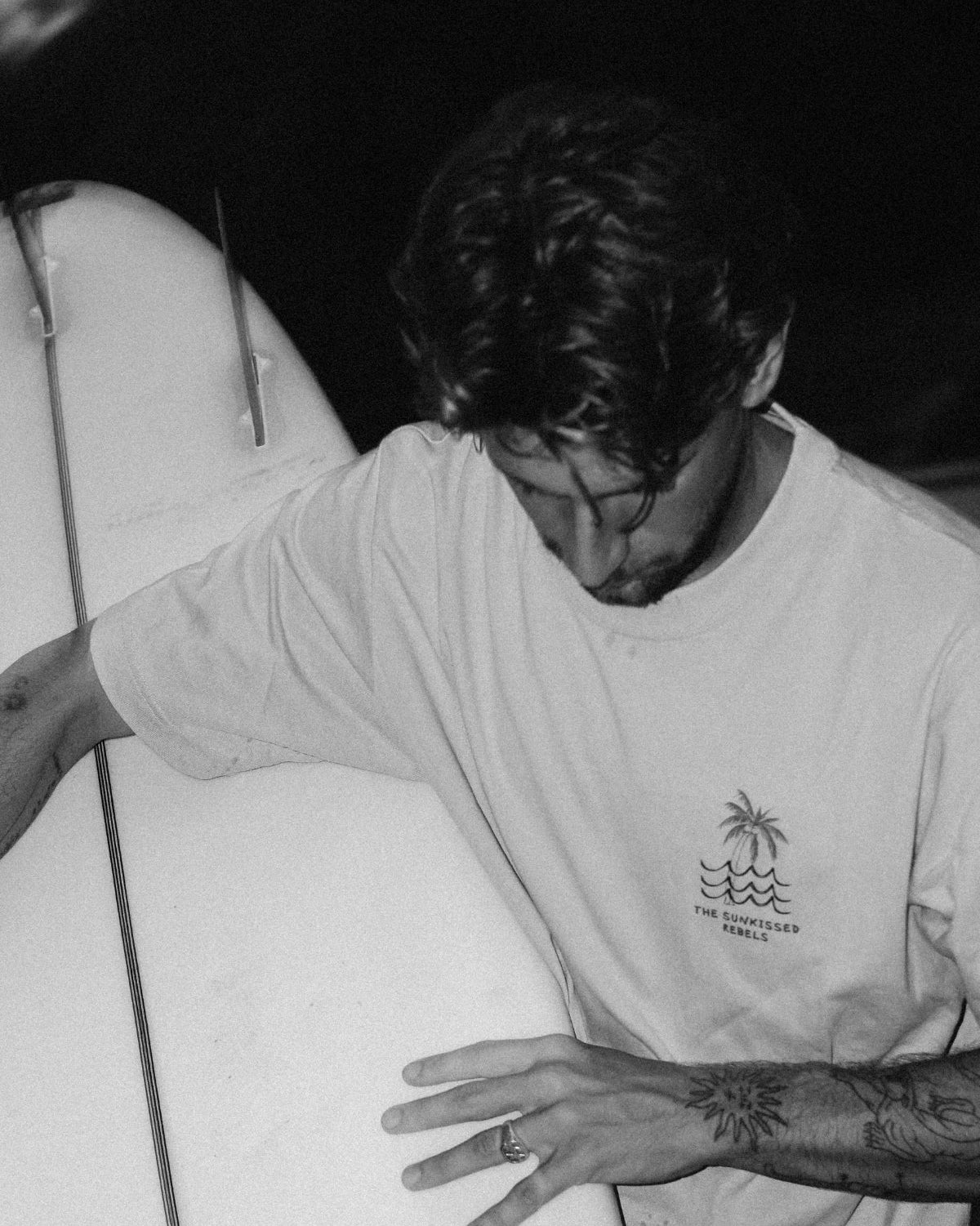 Schwarz-Weiß-Aufnahme eines Surfers mit dem Waves & Palm Trees Oversized T-Shirt in Sand und Surfboard am Strand.