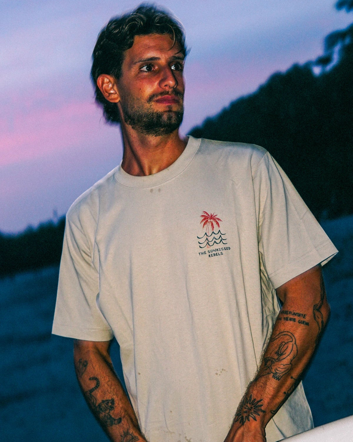 Mann trägt das Waves & Palm Trees Oversized T-Shirt in Sand mit Brustprint und Surfboard bei Sonnenuntergang am Strand.