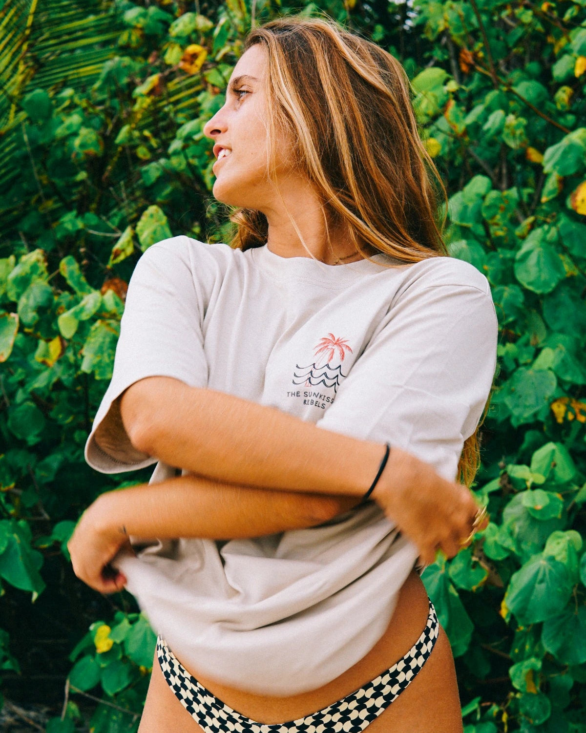 Frau trägt das Waves & Palm Trees Oversized T-Shirt in Sand vor tropischer Strandkulisse mit Palmen und grüner Vegetation.