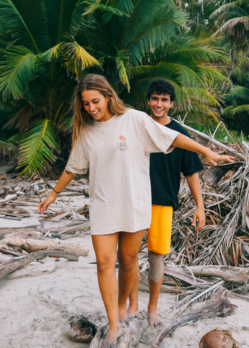 Surfer trägt das weiße Waves & Palm Trees Oversized T-Shirt von The Sunkissed Rebels mit Surfboard in einer stimmungsvollen Schwarz-Weiß-Aufnahme am Strand.