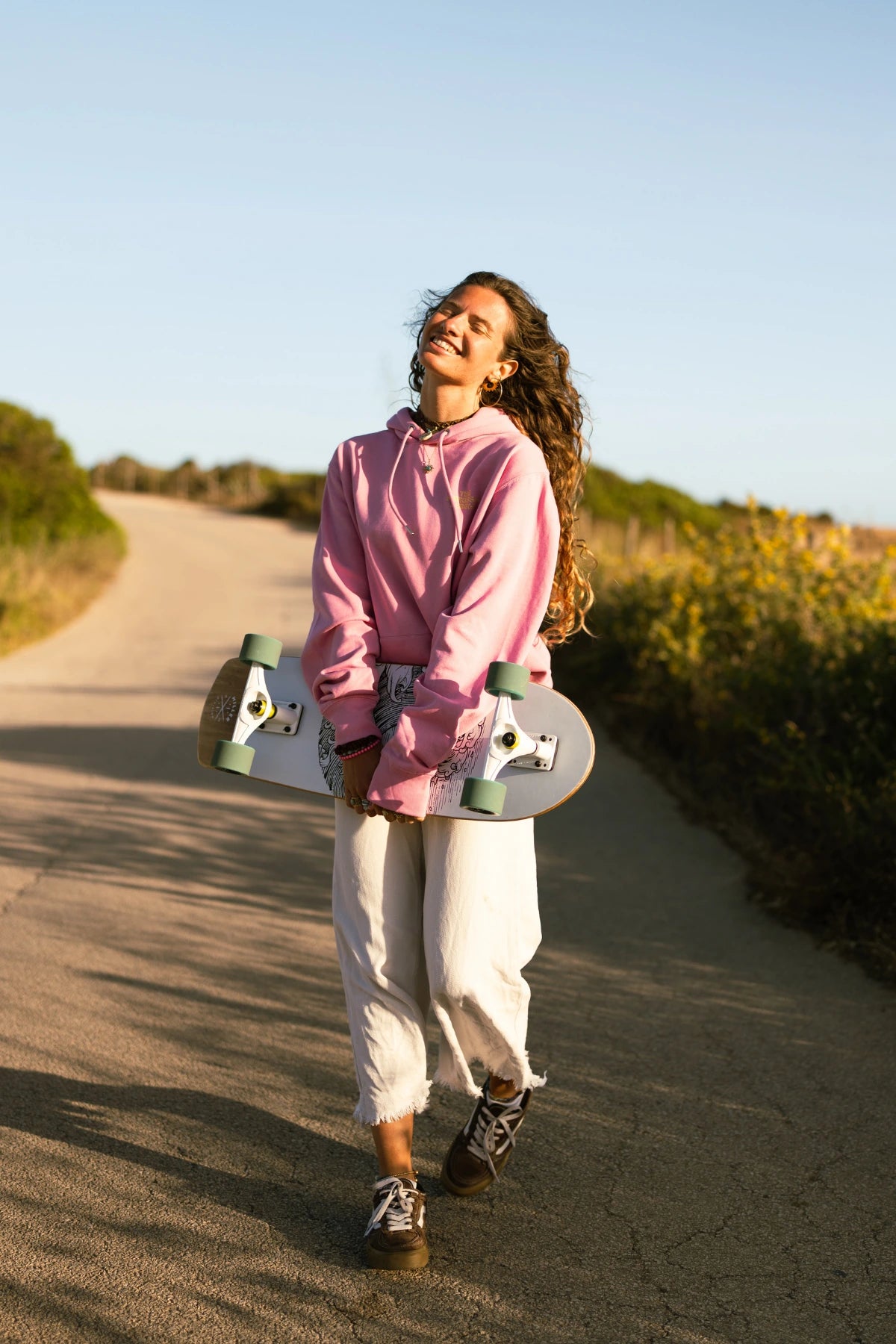 Frau trägt den A Good Day Hoodie Bubble Pink und läuft mit einem Skateboard auf einem sonnigen Küstenweg.