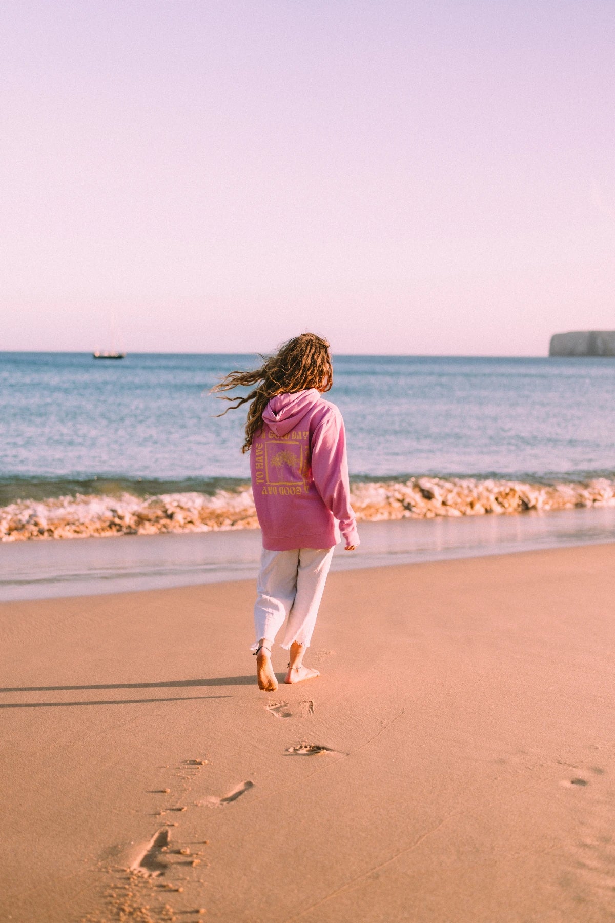 Frau im pinken A Good Day Hoodie spaziert barfuß am Meer entlang und hinterlässt Fußspuren im Sand.