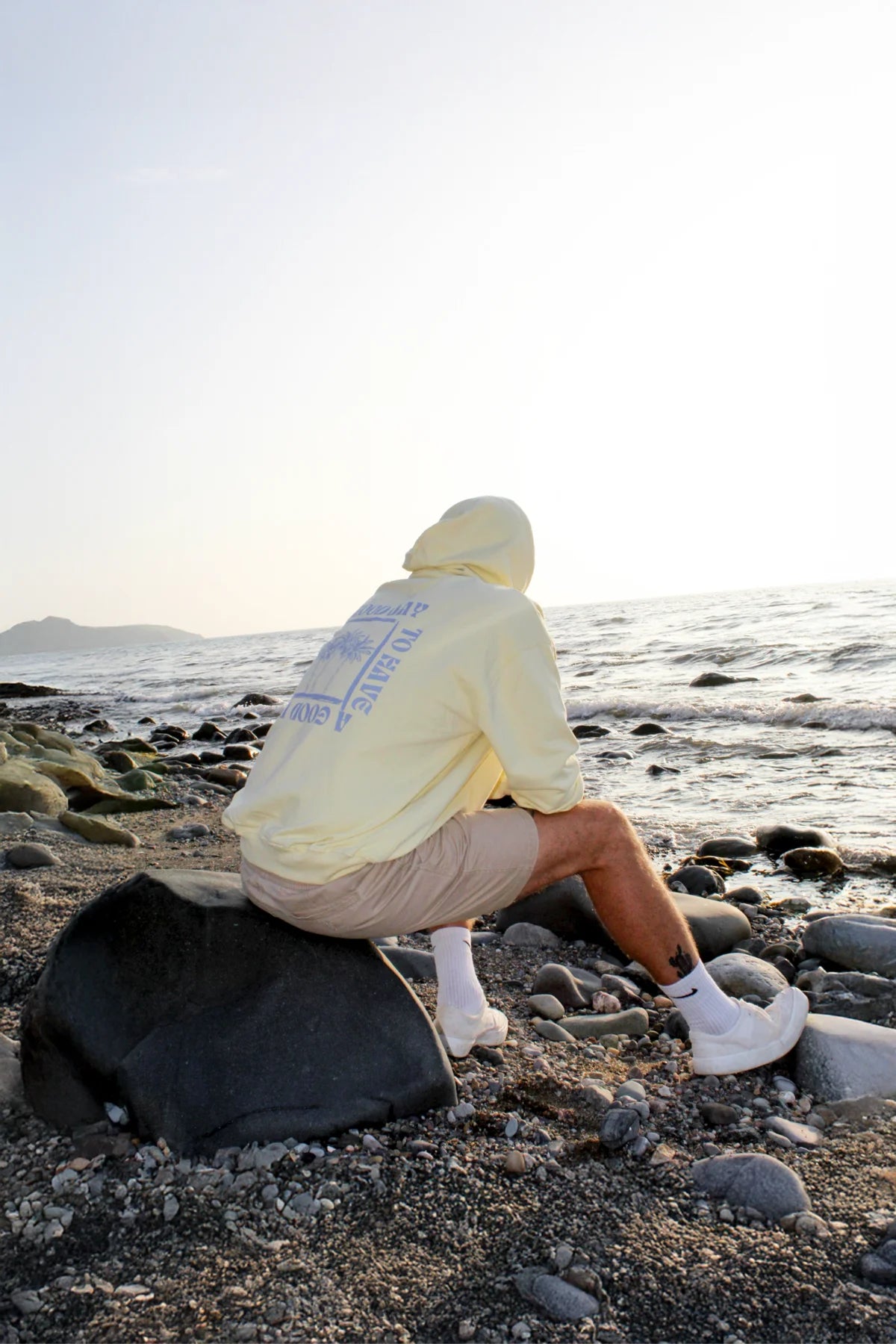 Person sitzt mit Hoodie auf einem Stein am Wasser, Sonnenuntergang reflektiert im Meer.