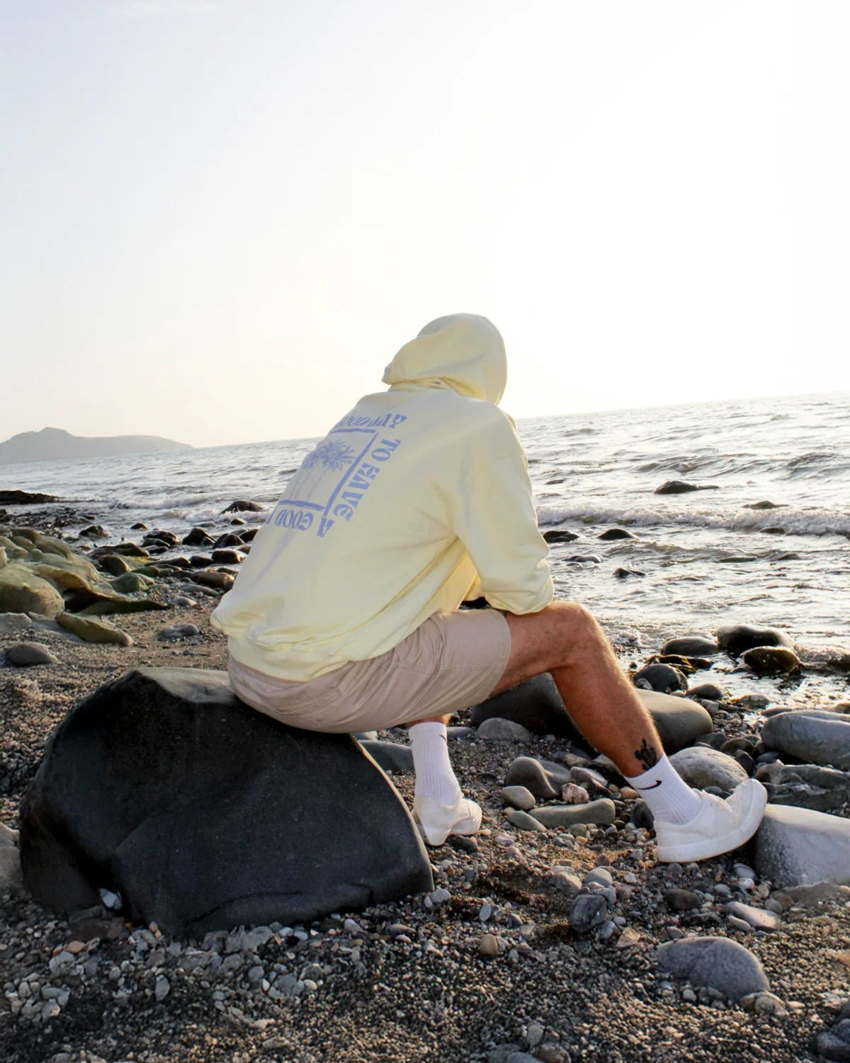 Person sitzt mit Hoodie auf einem Stein am Wasser, Sonnenuntergang reflektiert im Meer.