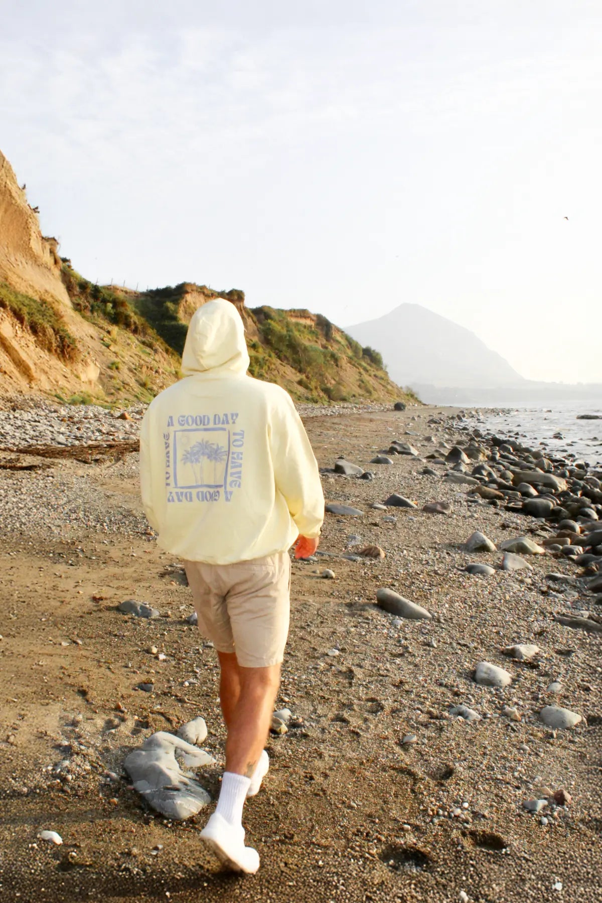 Person in buttergelbem Hoodie beim Spaziergang entlang eines felsigen Strands mit Bergen im Hintergrund.
