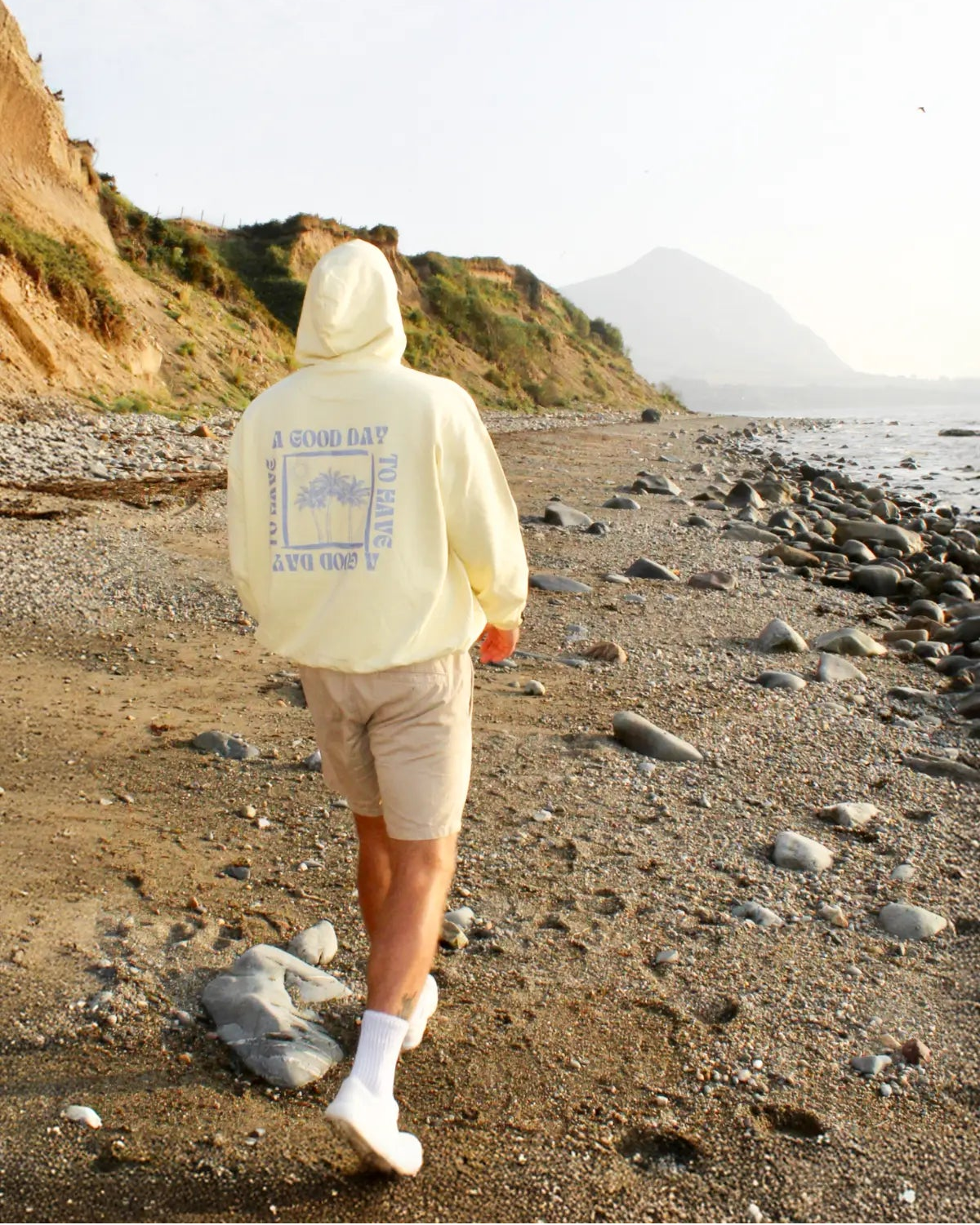 Person in buttergelbem Hoodie beim Spaziergang entlang eines felsigen Strands mit Bergen im Hintergrund.