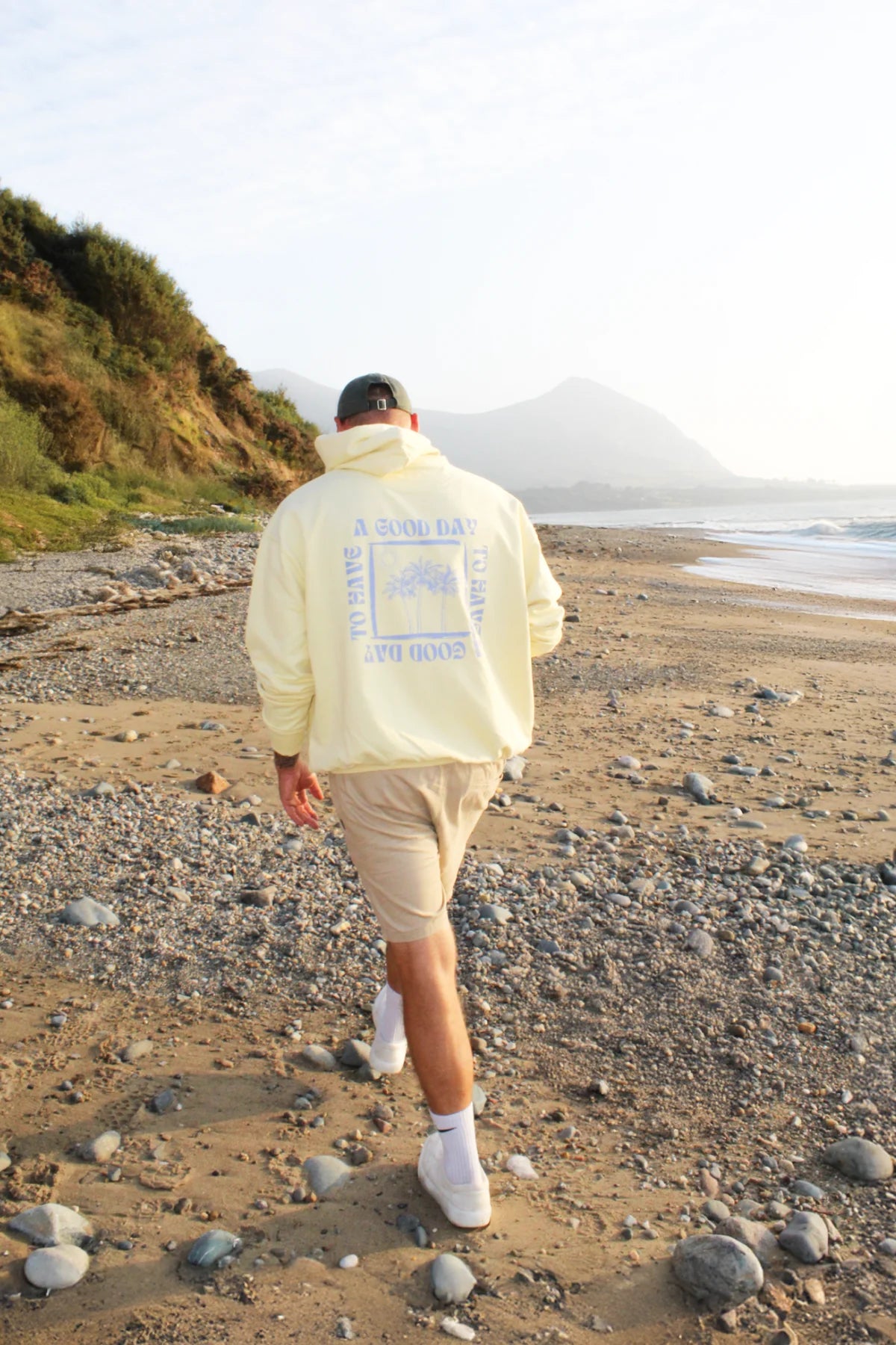 Rückenansicht eines Spaziergangs in butterfarbenem Hoodie entlang eines sonnigen Strandes.